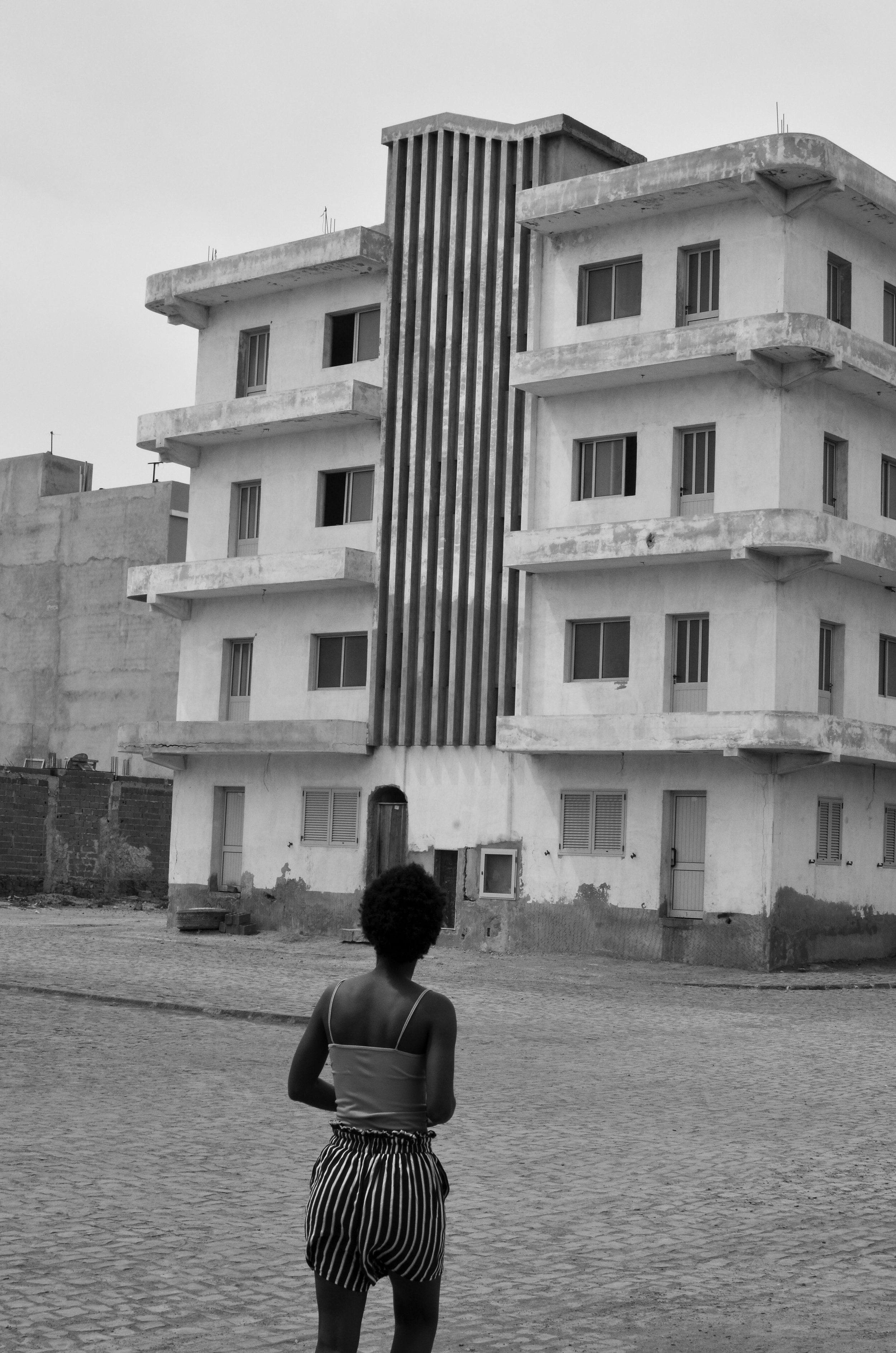 A young girl with short hair and striped shorts standing on a cobblestone street, facing a partially constructed or dilapidated multi-story building with a large vertical metal structure in front.