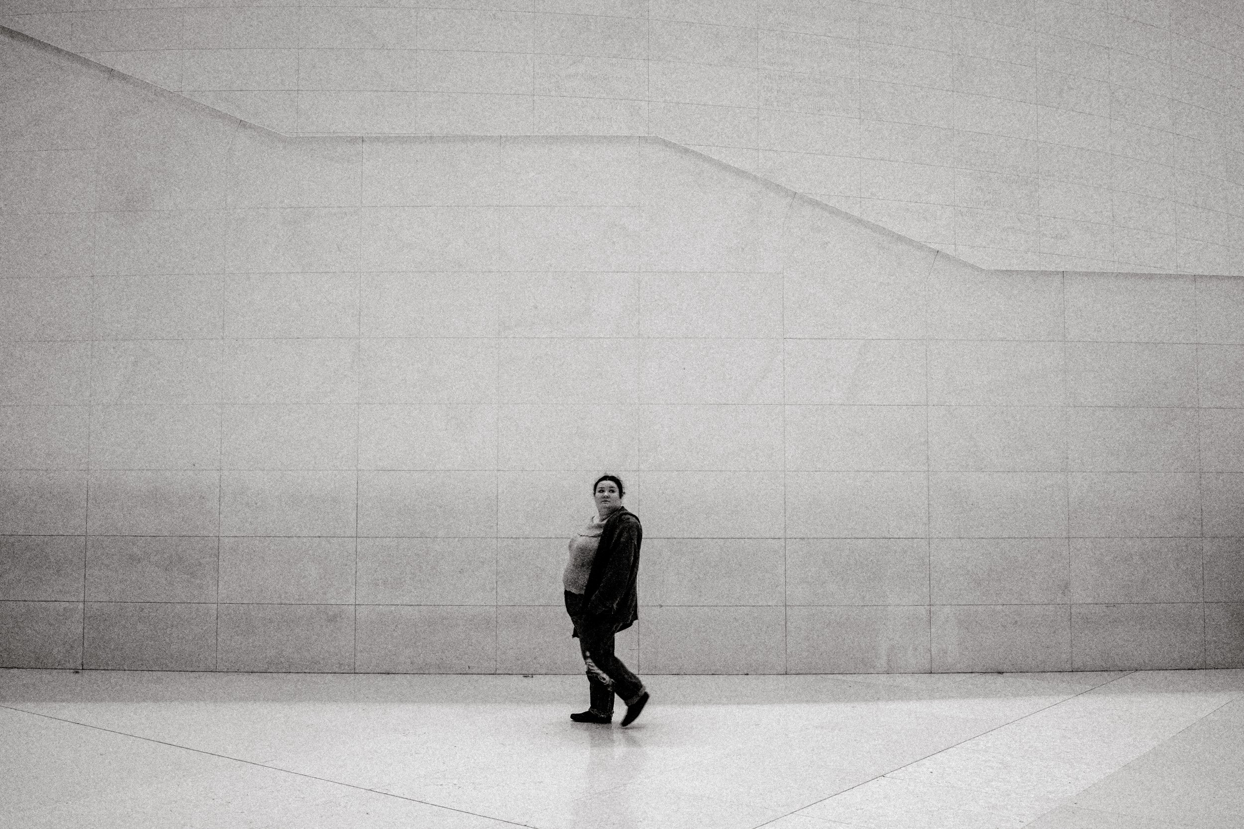 A woman in a dark jacket walks against a large, plain concrete wall in an indoor space