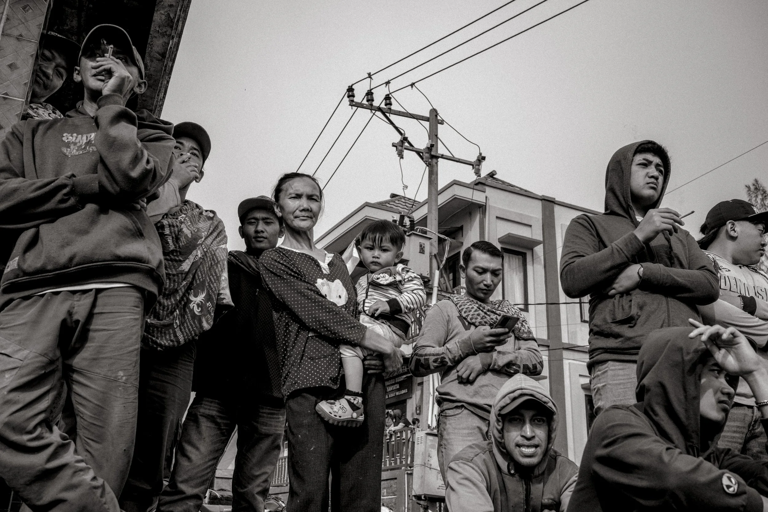 A group of diverse people standing outdoors, some with their arms crossed, others using a phone or holding children, with buildings and power lines in the background, all captured in black and white.