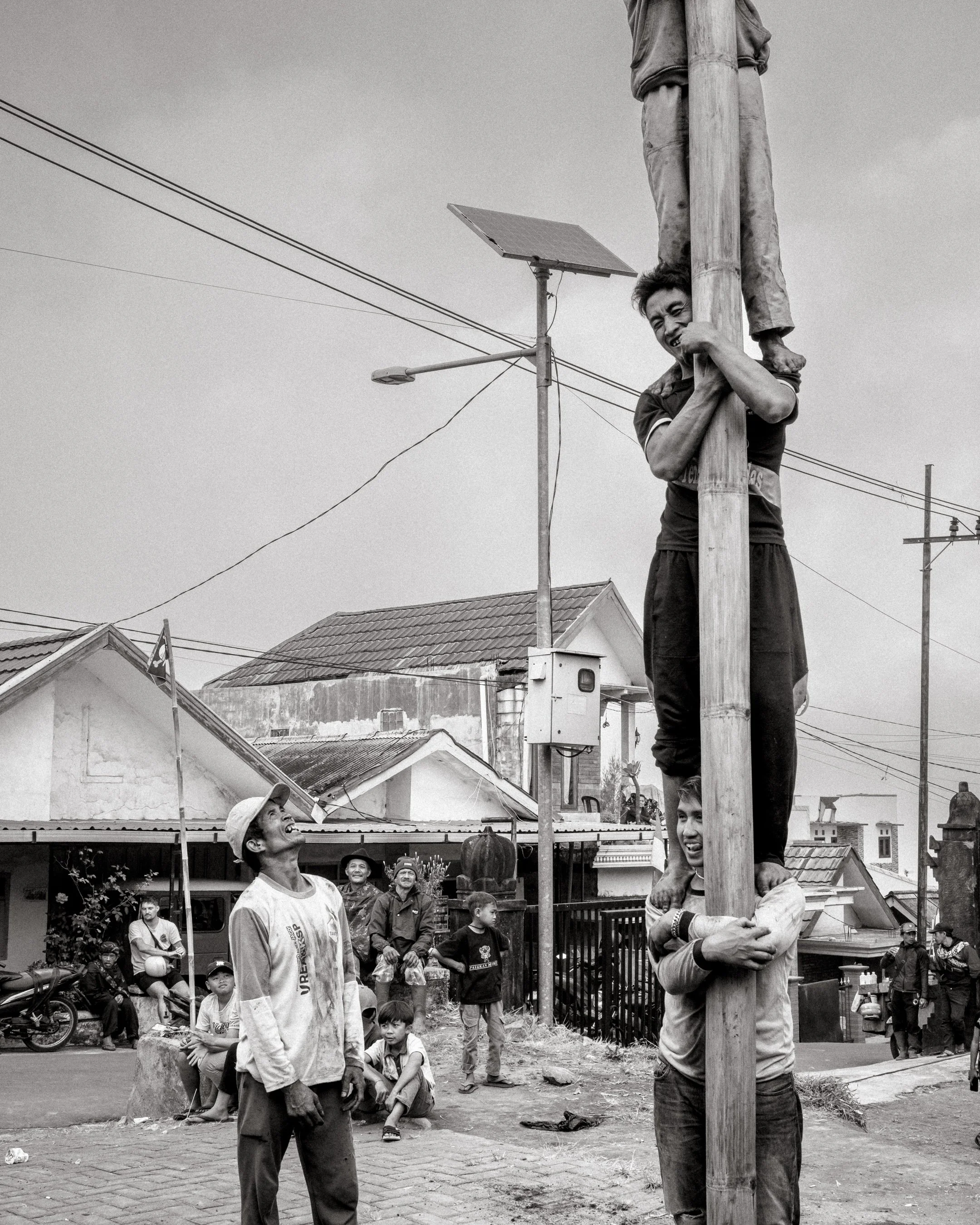 A team begins climbing as spectators watch from the street. Several climbers grin as they gather at the base. Ngadas, East Java, Indonesia. 21 August 2025.