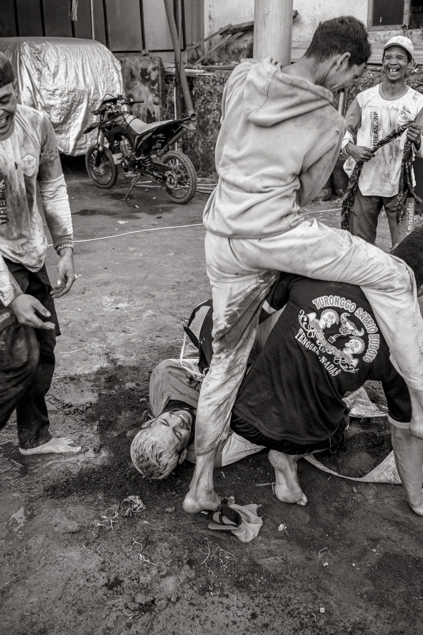 Climbers slip from the pole and fall into the mud, laughing. Participants wear a mix of local cultural group shirts and informal team prints rather than a single uniform. Ngadas, East Java, Indonesia. 21 August 2025.