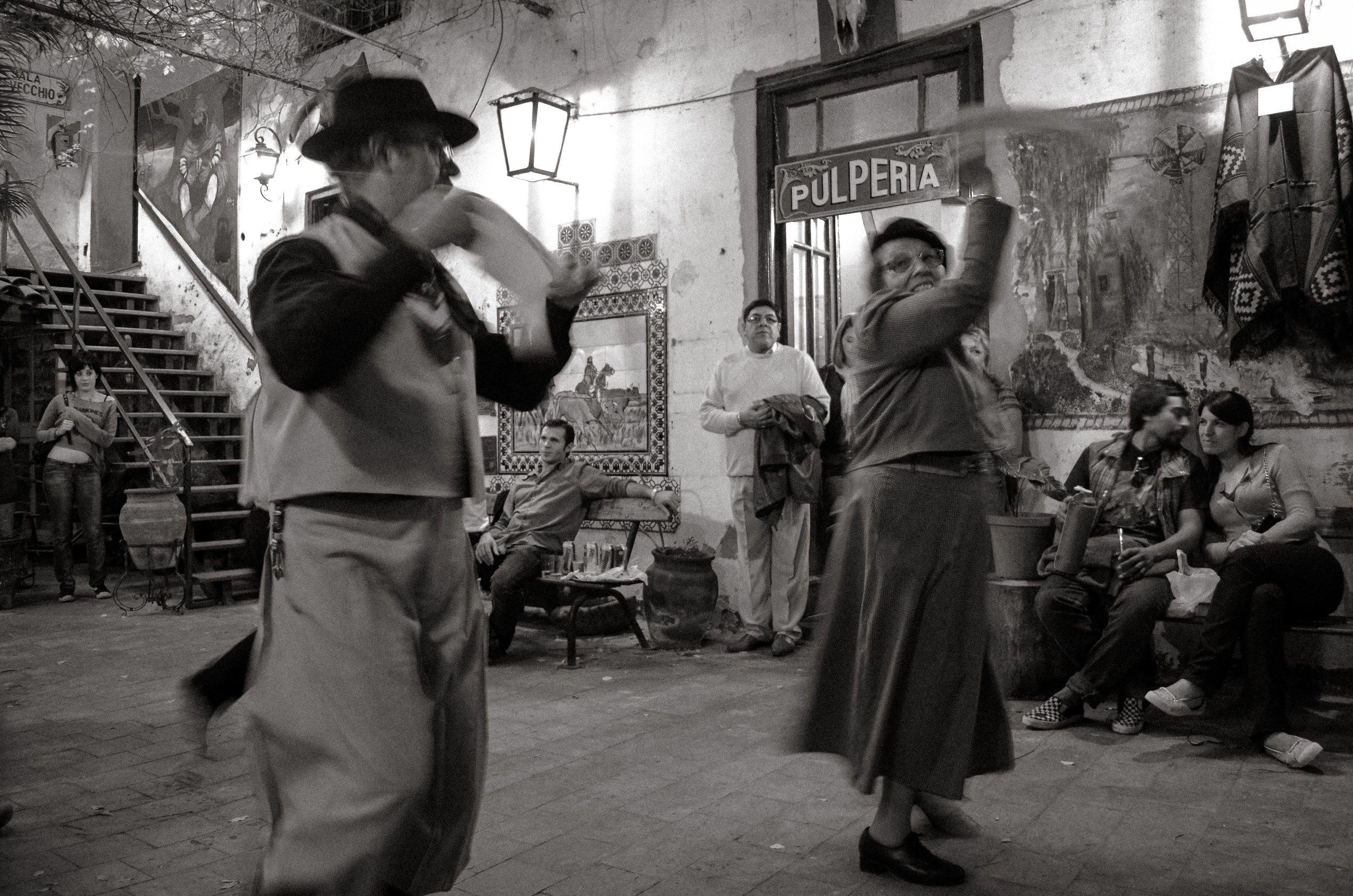 Two women dancing in a dimly lit, rustic indoor space with vintage decor, while several onlookers watch and sit on benches.