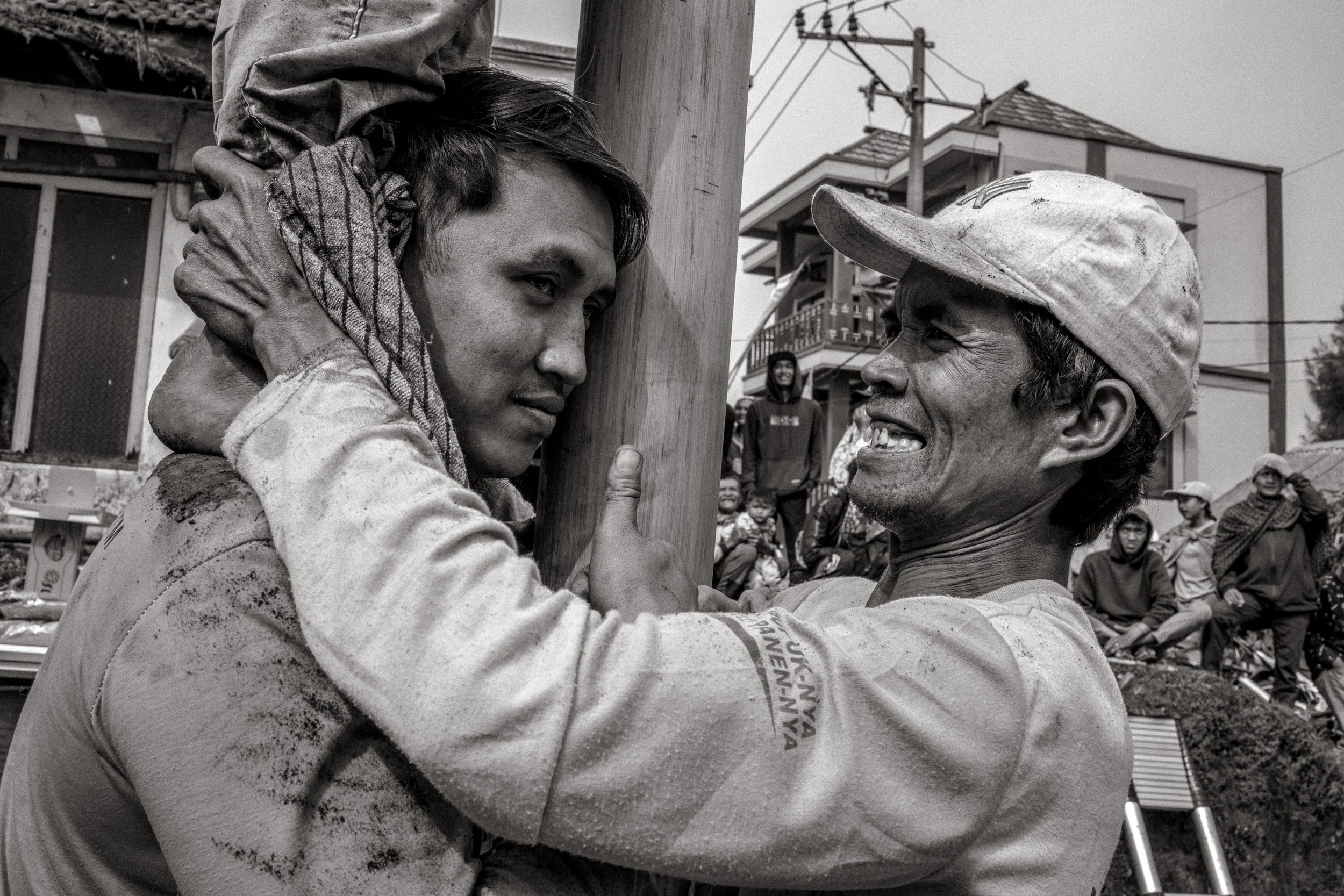 Two competitors brace the pole and grip each other to support the base as teammates climb above them. Behind them, spectators watch from the square. Ngadas, East Java, Indonesia. 21 August 2025.