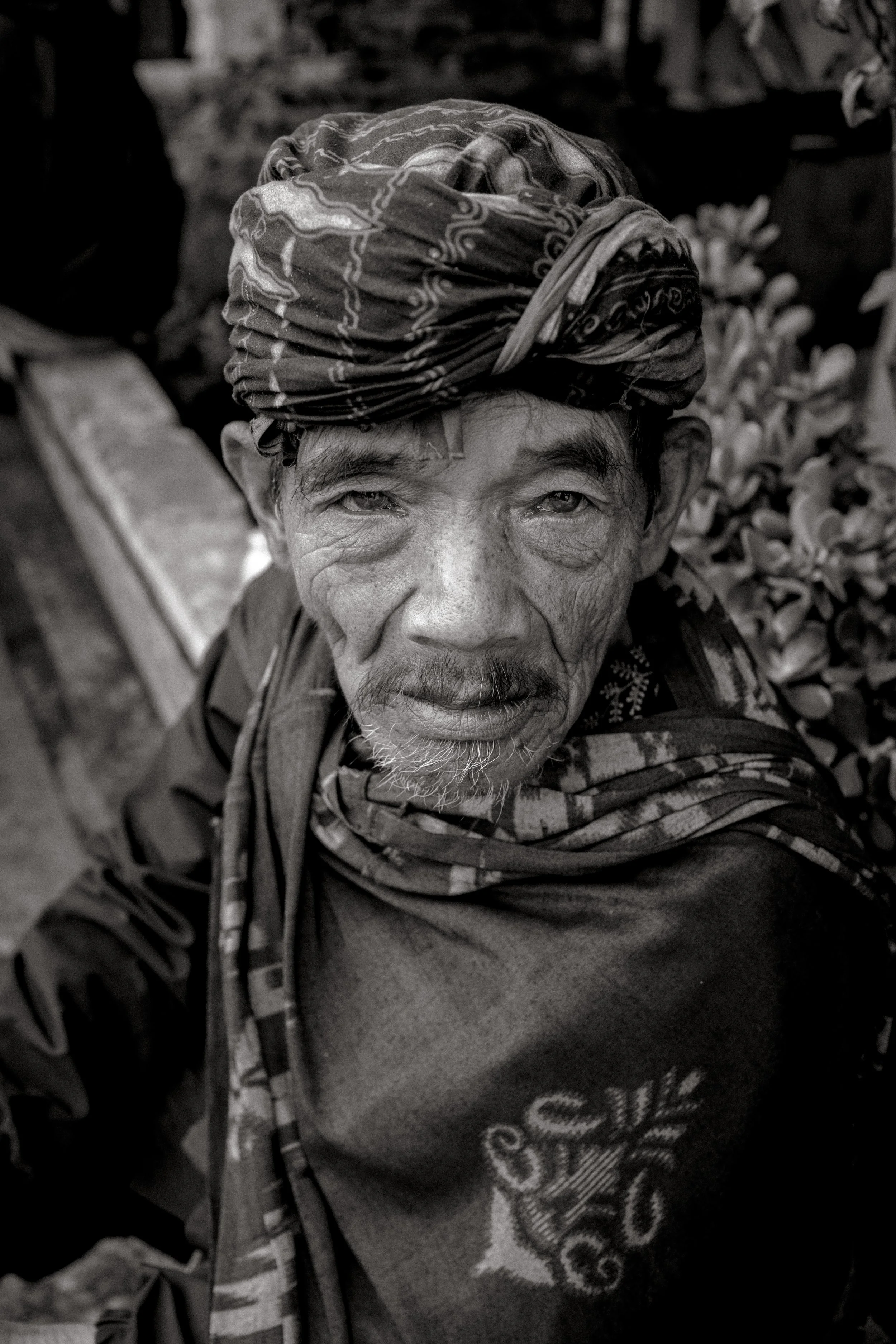 Black and white close-up of an elderly man wearing a traditional headscarf, with a confident expression, snowy beard, and textured skin, outdoors with plants in the background.