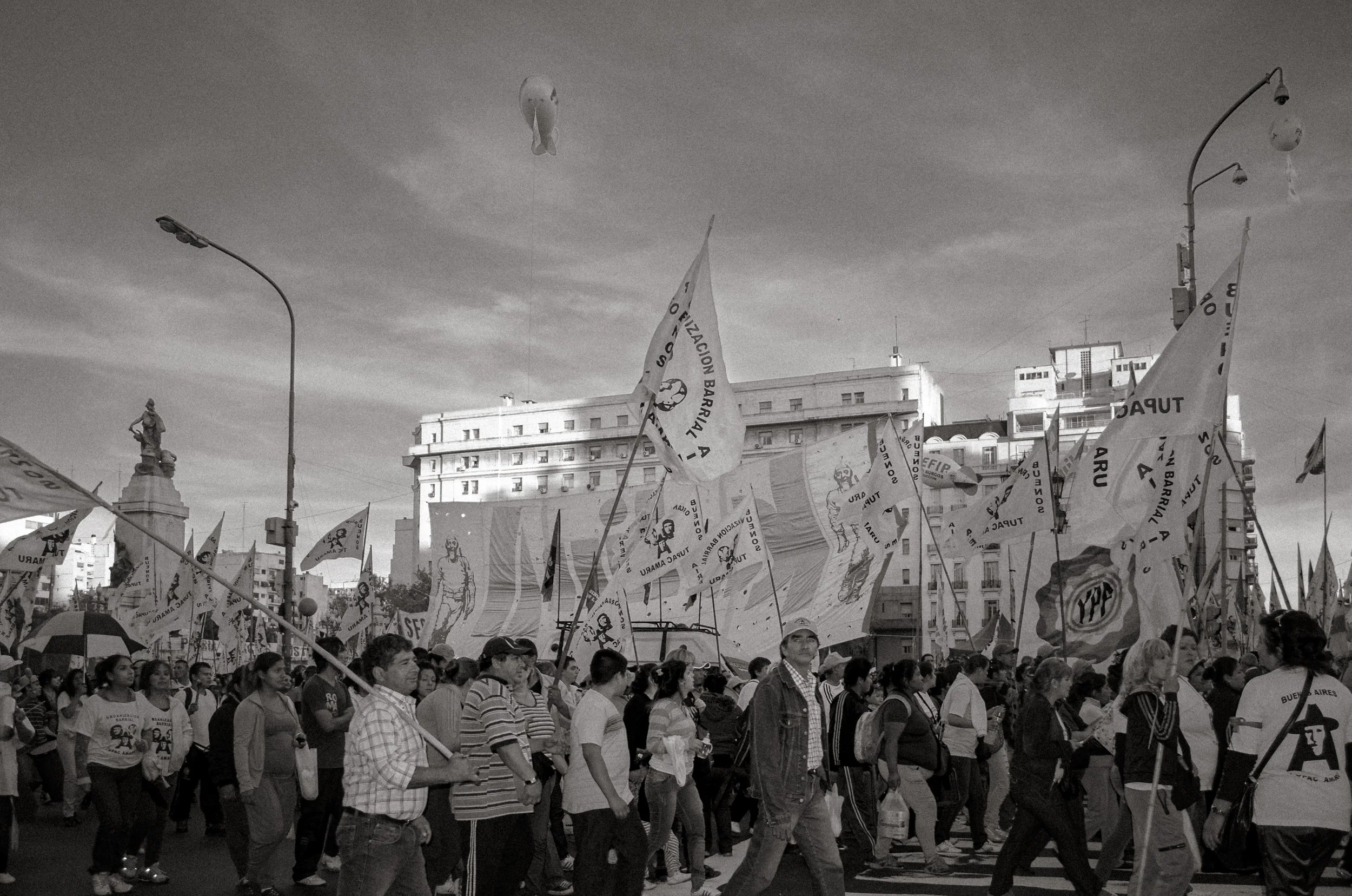A large crowd of people participating in a march, carrying numerous flags with symbols and text, walking through an urban area with tall buildings and a statue in the background; the scene is under a cloudy sky.