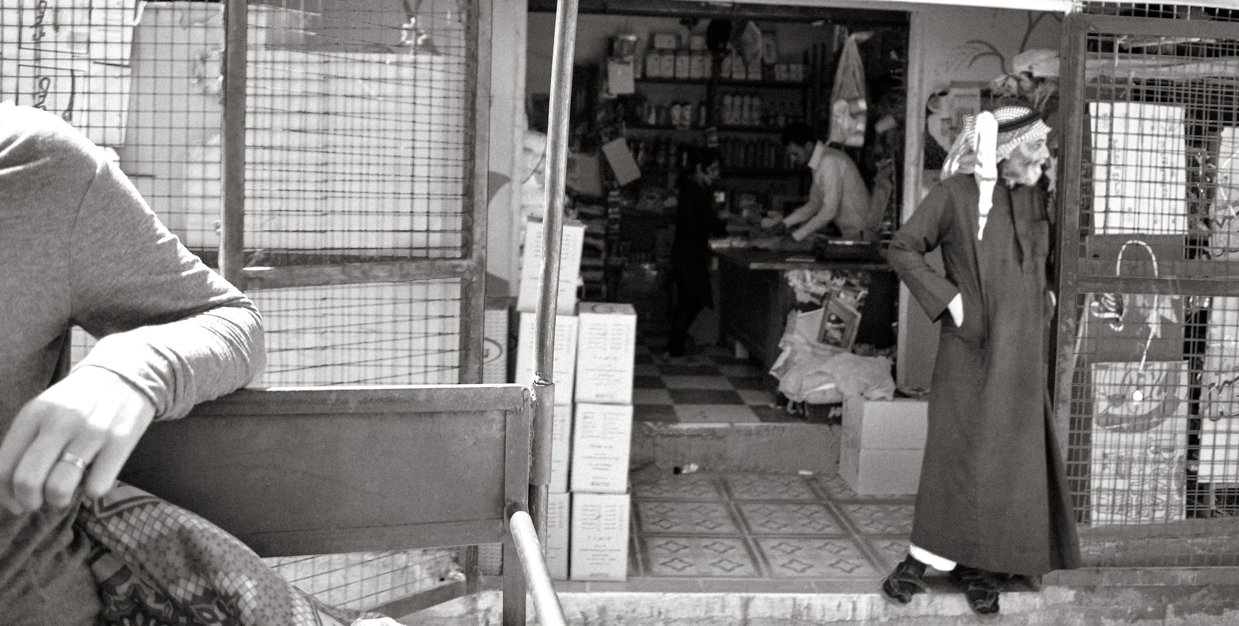 A man in traditional Middle Eastern attire standing outside a small shop, with a woman working inside behind the counter and another person visible inside. The shop has shelves filled with various products and is enclosed by wire fencing.