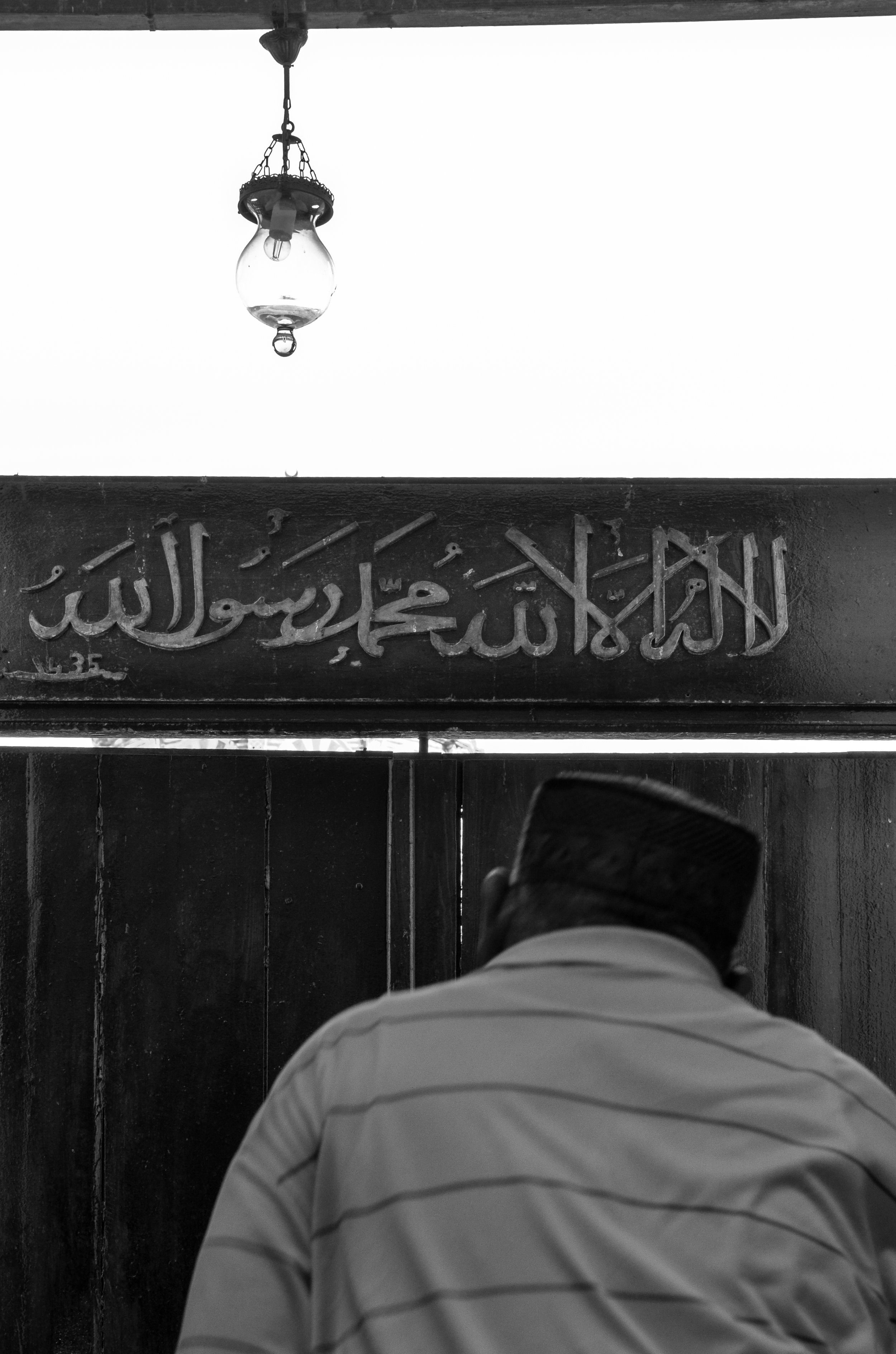 A man wearing a cap is sitting with his back facing the camera. Above him is a sign with writing in Arabic script. There is a hanging light fixture above the sign. The photo is in black and white.