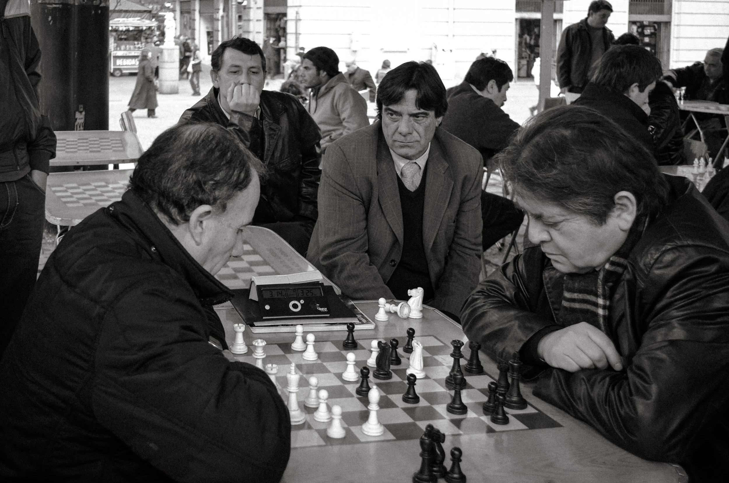 Two men playing chess outdoors in a busy plaza, with onlookers and more tables in the background.