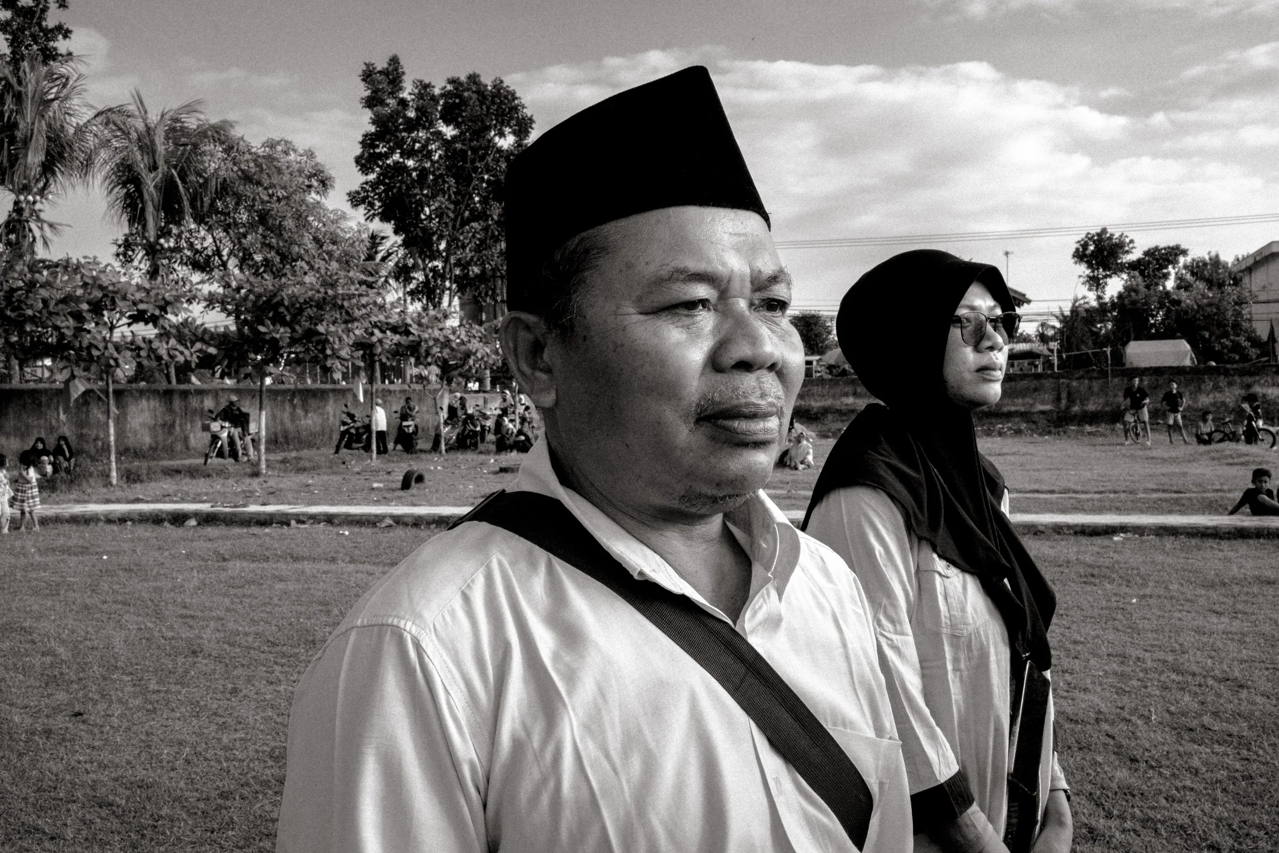 A black and white photo of a man wearing a traditional Indonesian cap (peci) and a woman wearing a hijab standing outdoors, with a park and people in the background.