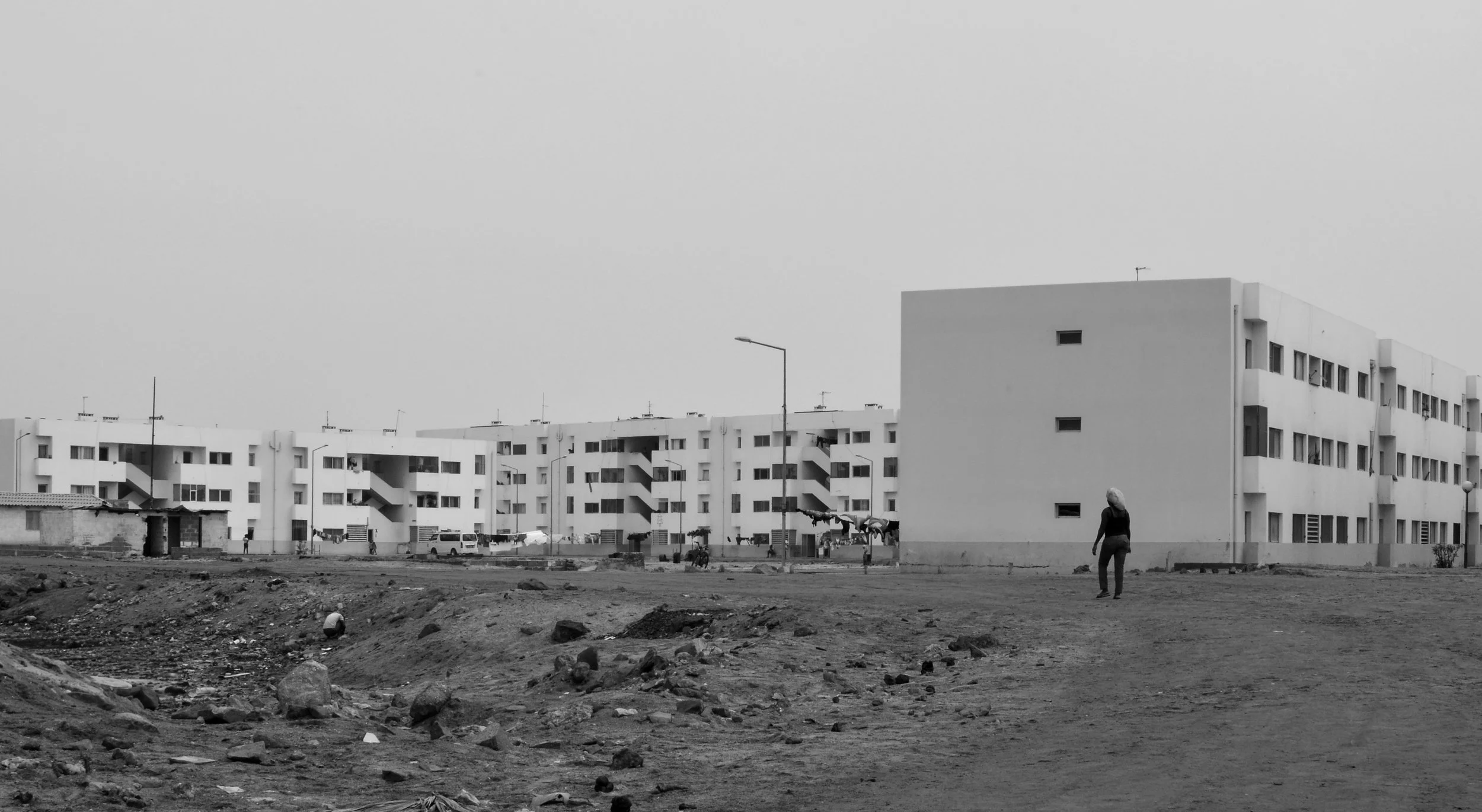A black and white photo of an urban area with multistory apartment buildings, a woman walking, and a muddy, rocky ground in the foreground.
