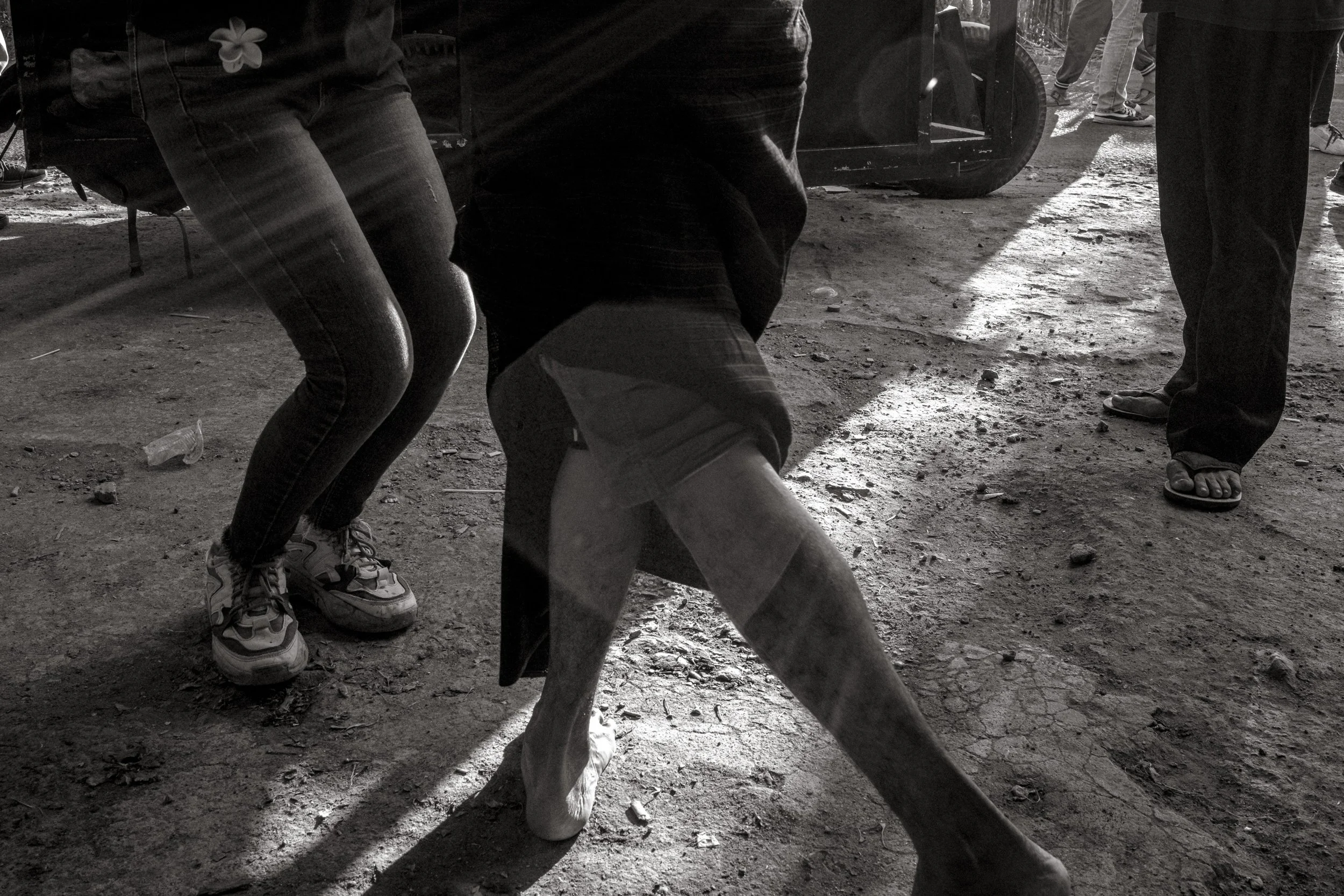Bare feet and trainers step across dust and mud in hard light as dancing continues