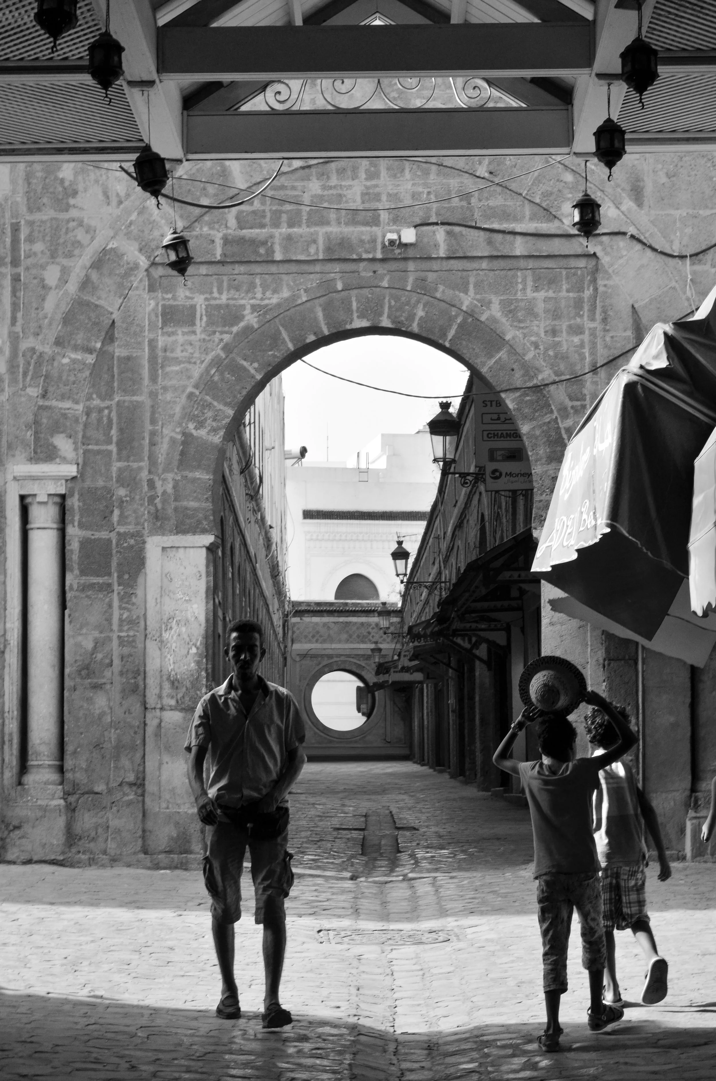 Black and white photo of a street scene with an arched stone gateway. Two children walk away from the camera, one holding a hat, with a man walking toward the camera. Buildings and lampposts line the street.