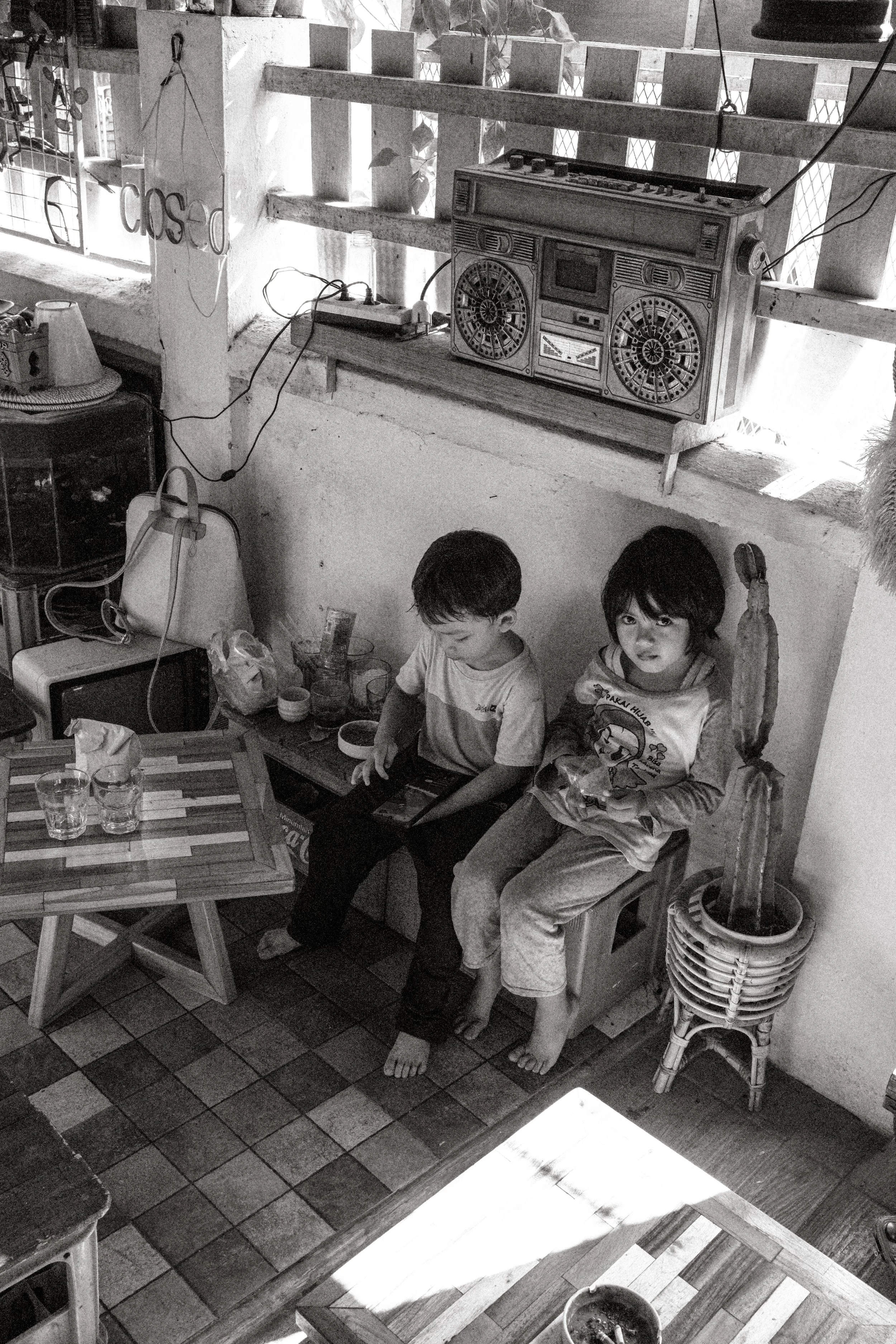 Two young boys sitting barefoot on a small wooden box and a chair, one using a tablet and the other holding a snack, in a cozy indoor space with a retro radio on a shelf and various household items around.