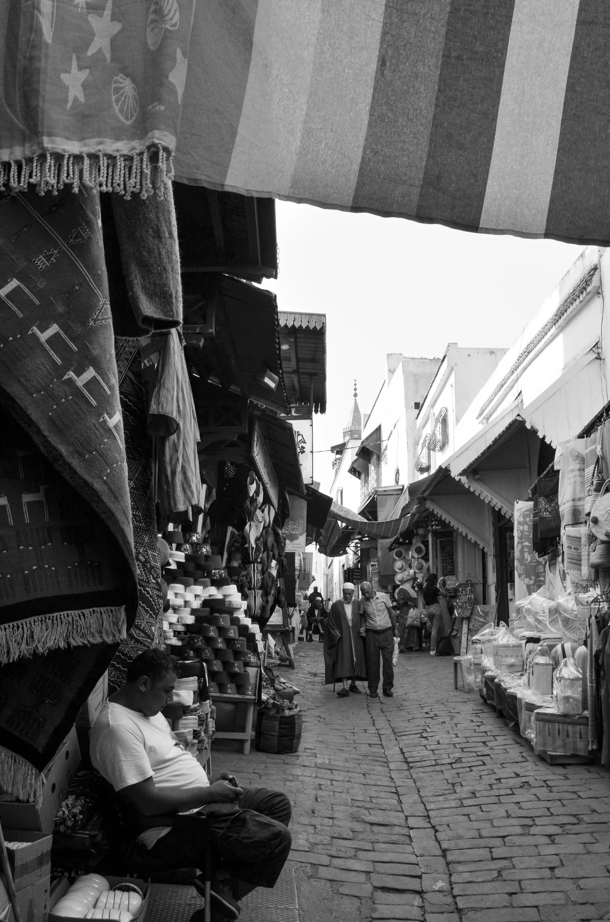 People walking and shopping in a busy outdoor marketplace with stalls selling textiles, hats, and other goods, cobblestone street, and a church steeple in the background.