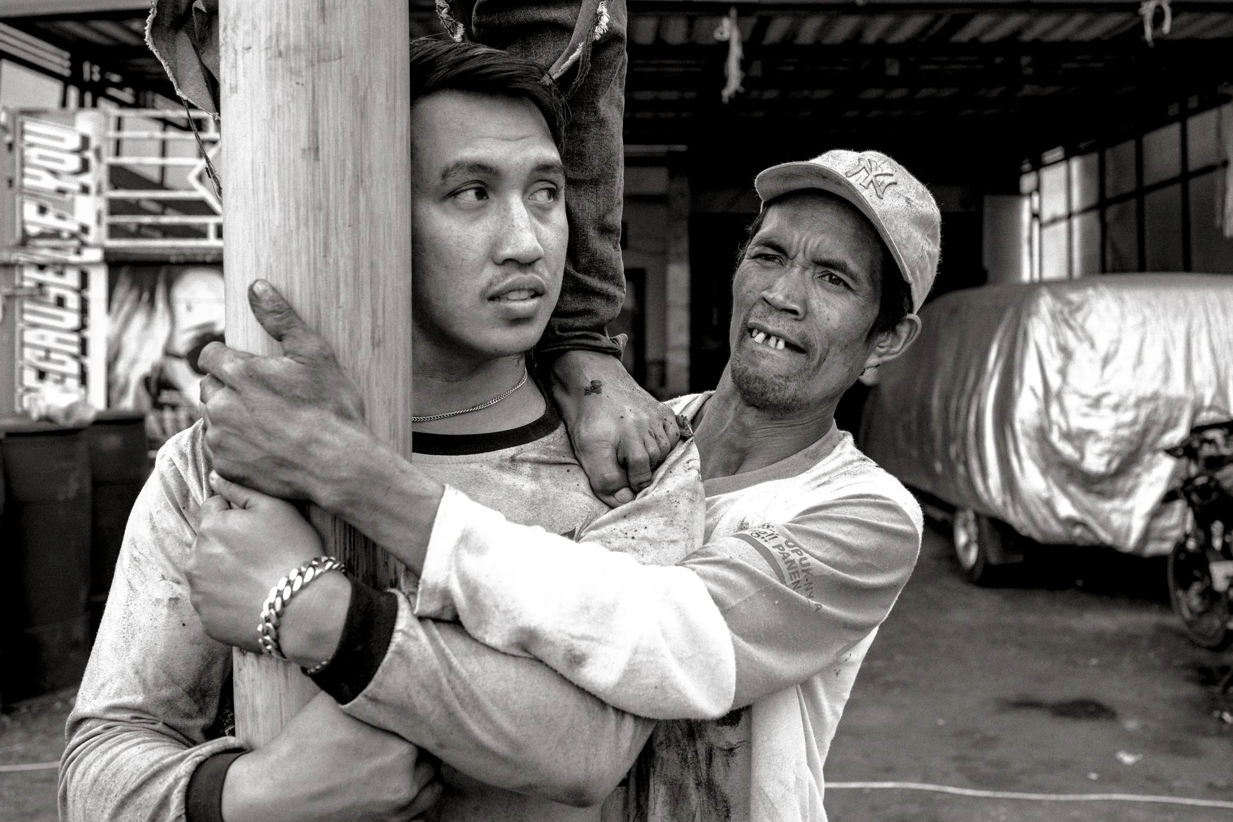 A black and white photo of a man holding a young boy, who is secured to a wooden post, with an expression of concern or distress. The scene appears to be in an industrial or market setting with a covered roof and large covered trucks in the backgroun