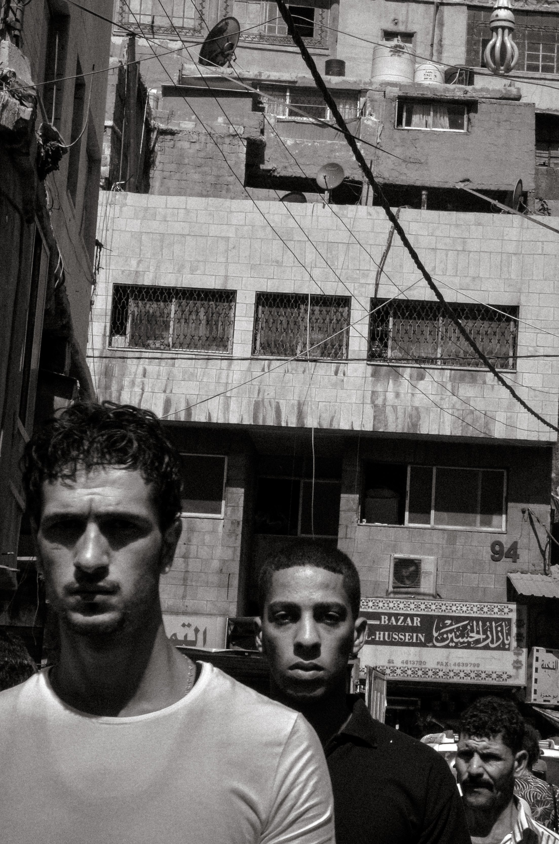 Black and white photo of three young men standing in a crowded street scene with urban background, buildings, wires, and signs.