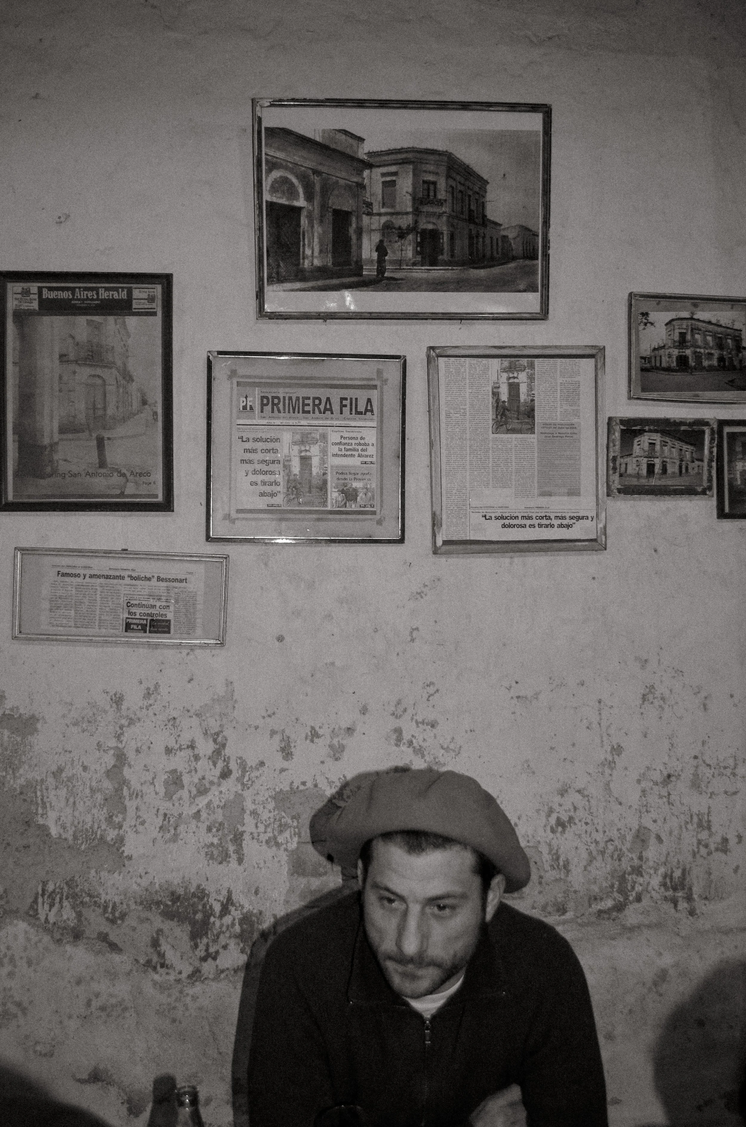 A man with a mustache and beard wearing a beret, sitting at a table in front of a wall with framed pictures and newspaper clippings, in a black-and-white photo.