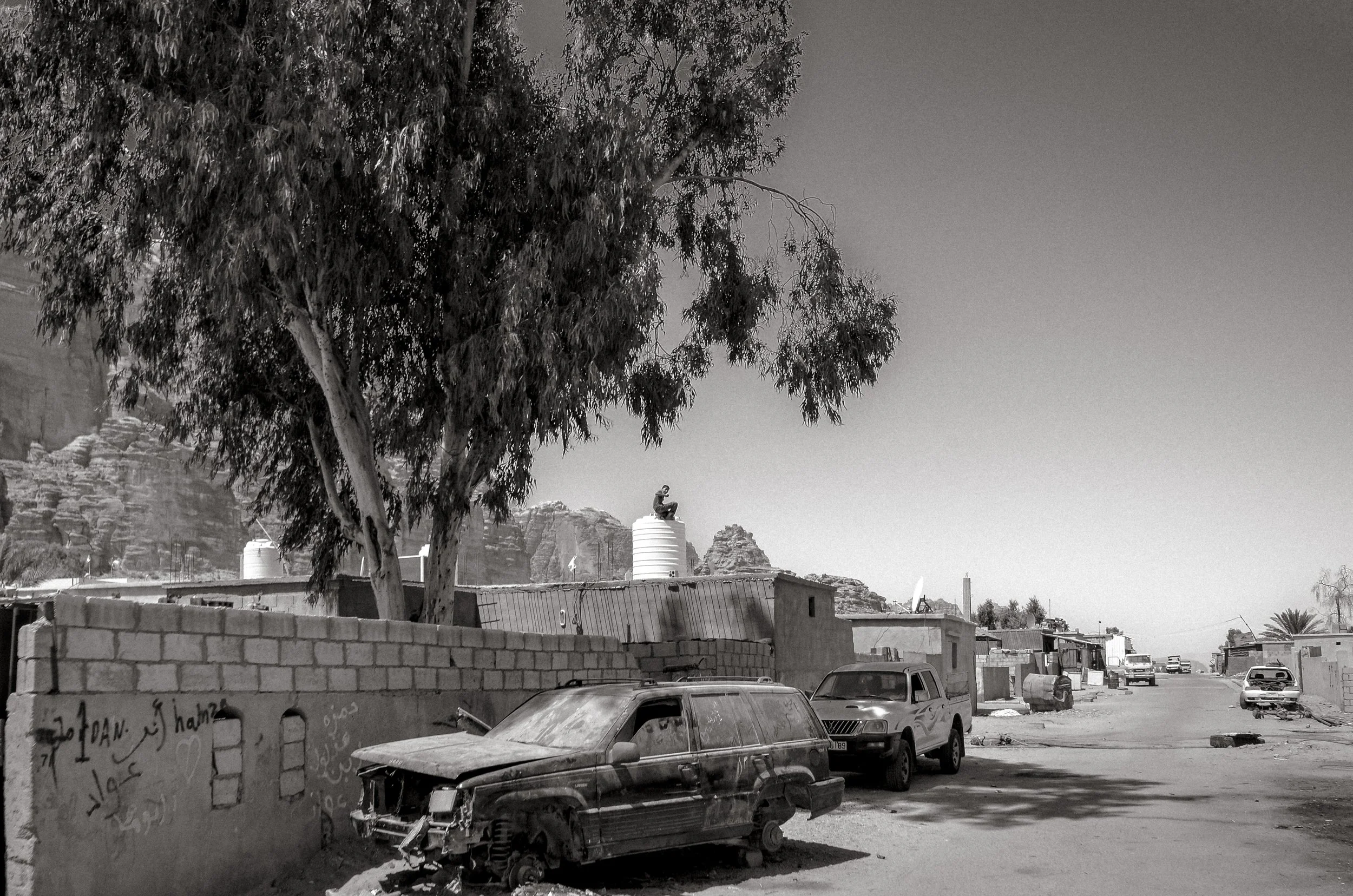 Black and white photo of a damaged car parked in a dusty street with other vehicles, a brick wall with writing, a large tree, and a background of rocky hills.