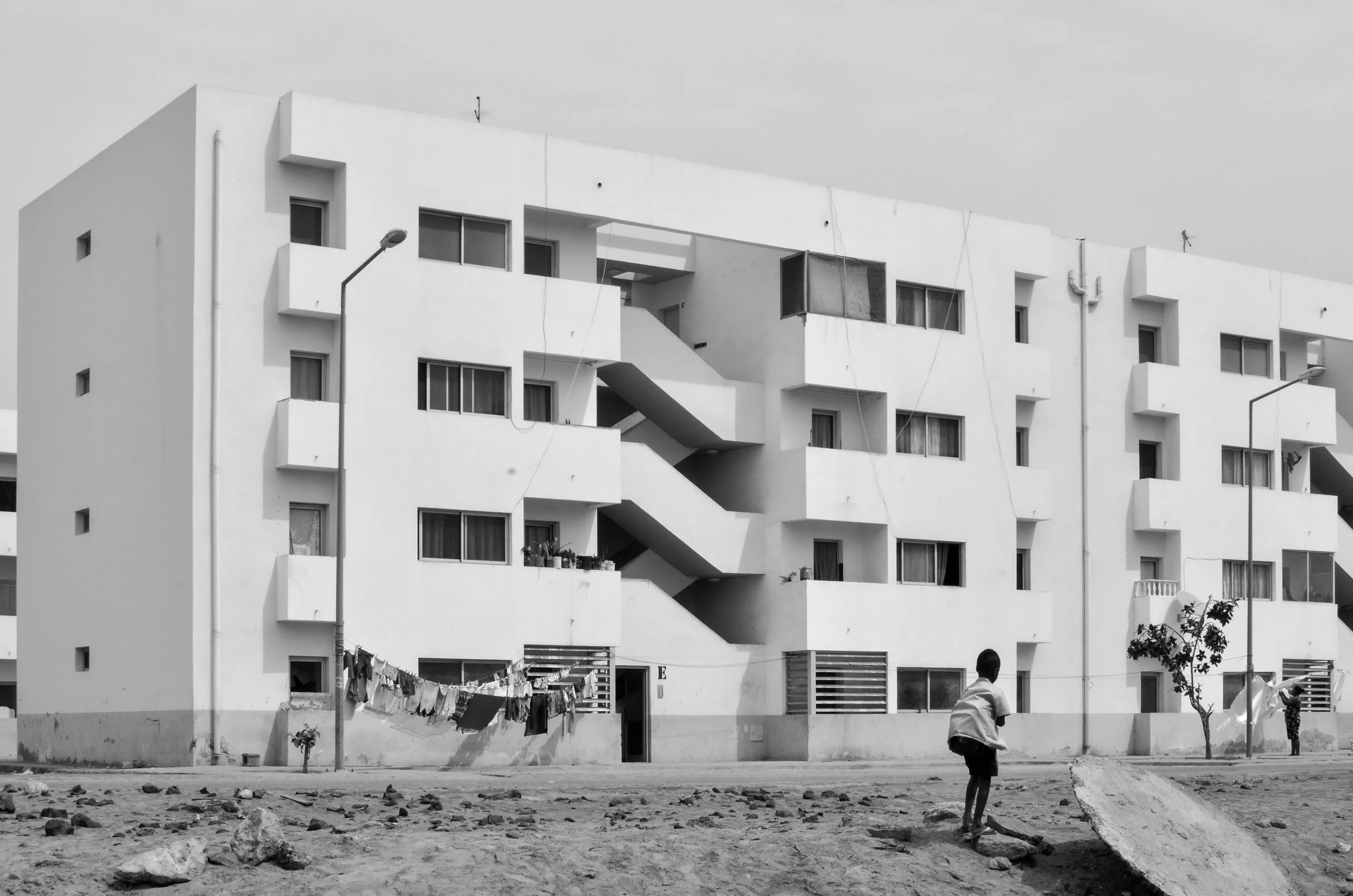 A black and white photo of a white apartment building with exterior staircases, surrounded by a dirt area with rocks. In the foreground, children are playing and laundry is hanging outside.