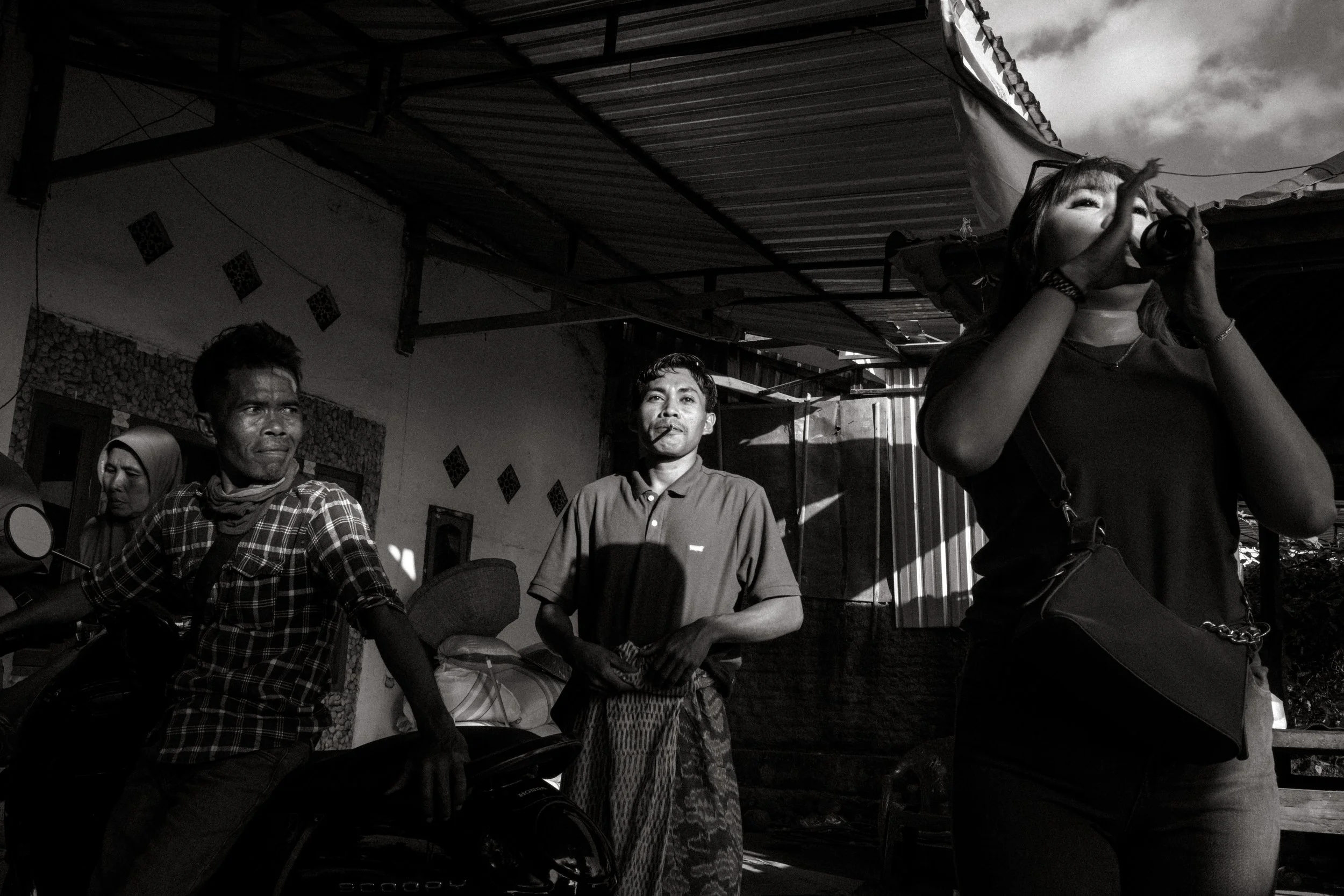 Black and white photo of three people in an outdoor setting. One woman is drinking from a bottle, two men are standing nearby, one with a cigarette in his mouth, and a woman in the background wearing a headscarf.