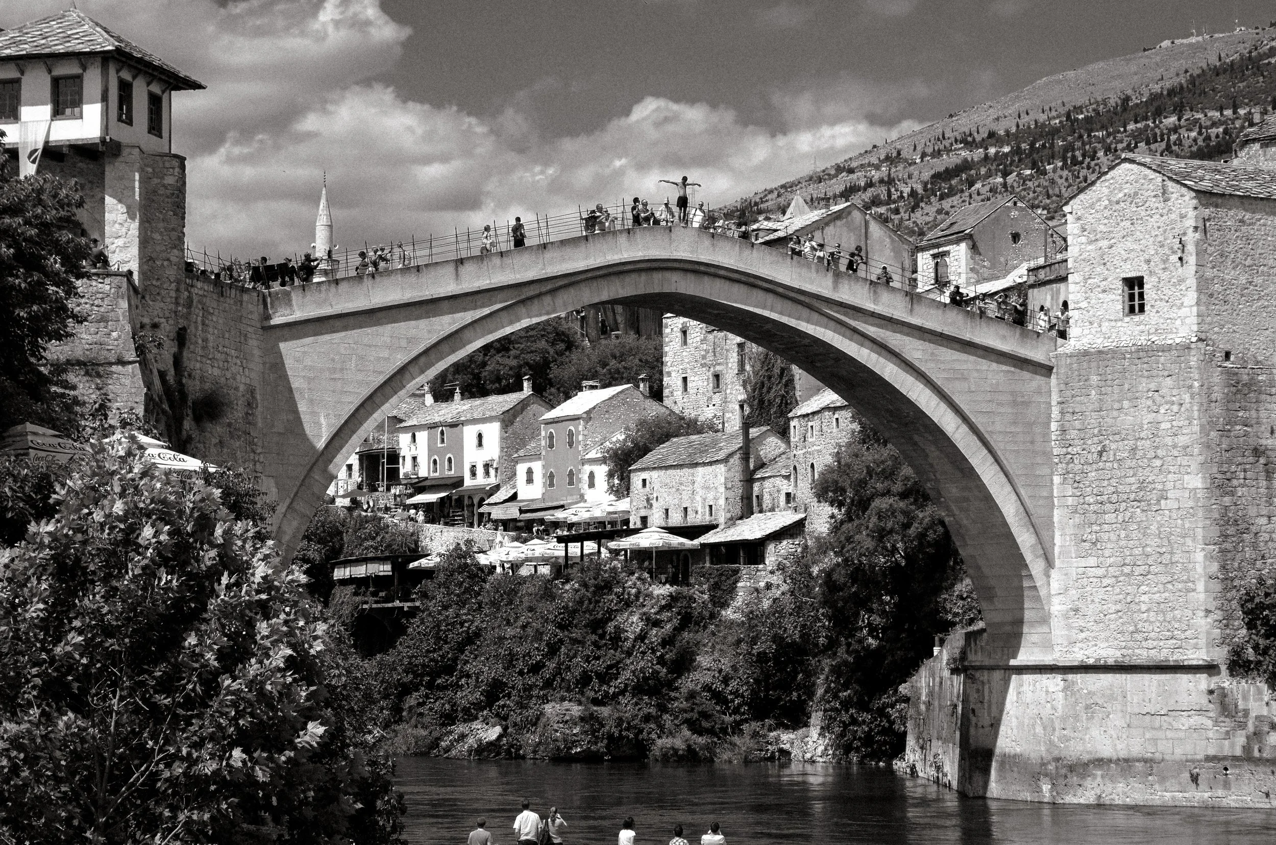 A stone arch bridge with people walking across from one side to the other, spanning over a river. The background features hillside houses, trees, and a cloudy sky in black and white.