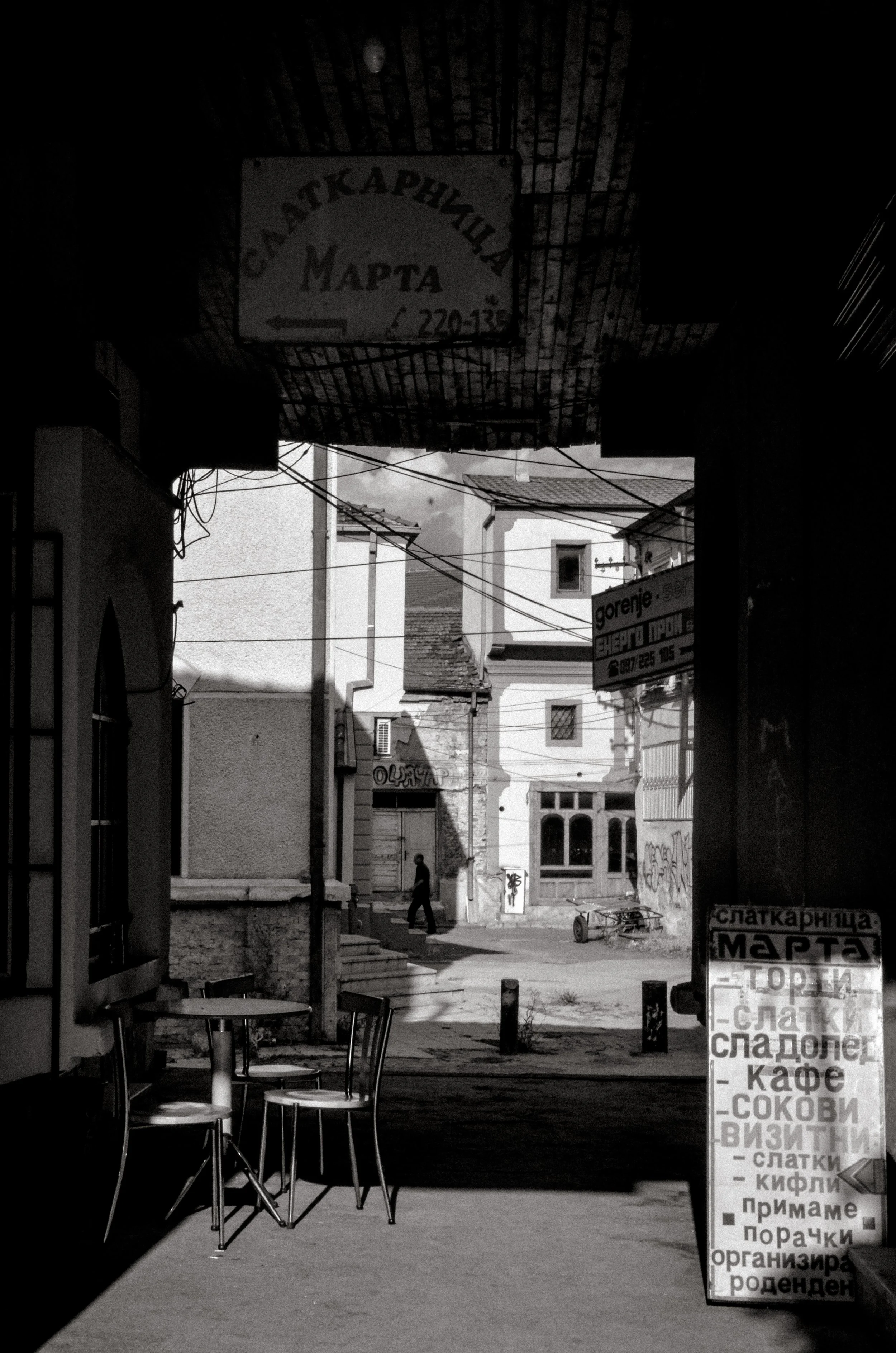 A black and white street scene viewed from inside a cafe or shop, with chairs and tables visible in the foreground. Outside, a person is walking down the street, with buildings and signs in various languages, including Cyrillic, visible in the backgr