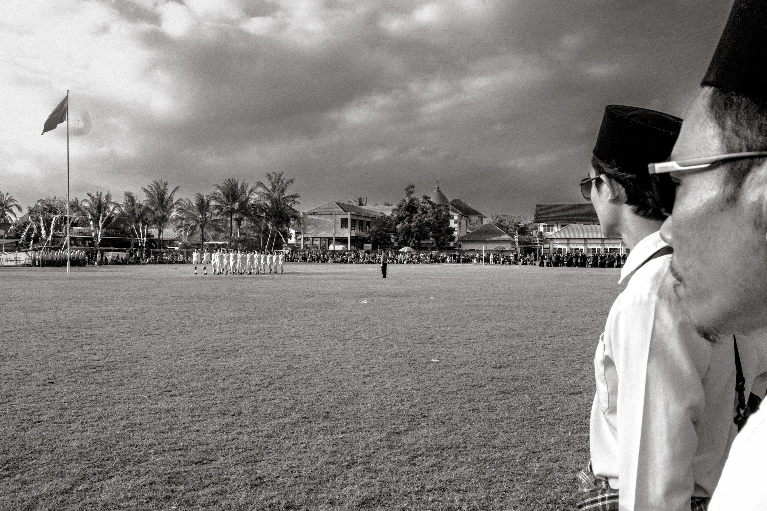 A black-and-white photo of a flagpole with a flag, students in white uniforms in a formation on a school field, some in traditional attire, and a crowd of spectators gathered behind a fence, with buildings and palm trees in the background.