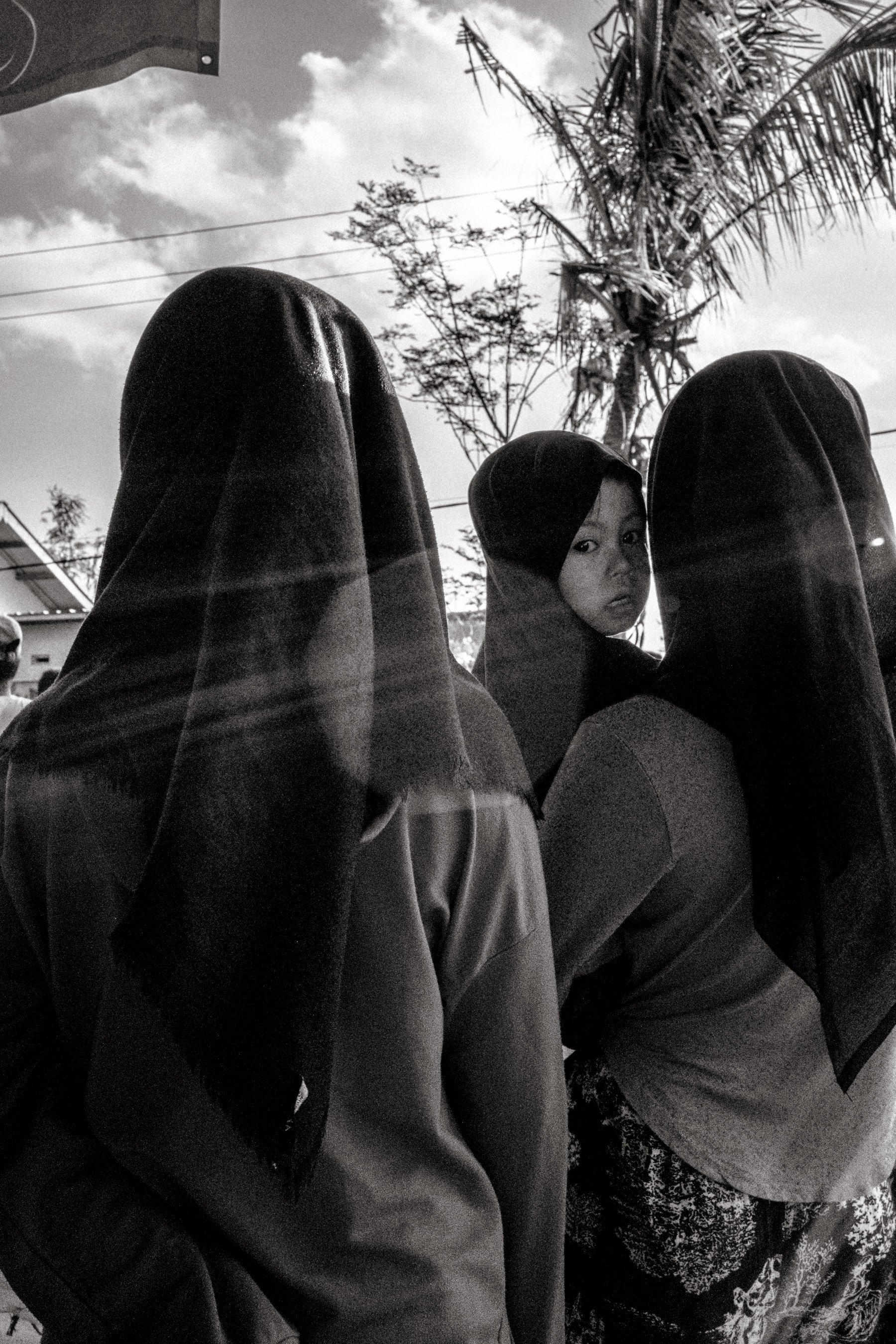 Women stand watching the event, as a small girl looks back. The Sasak are Lombok's largest ethnic group and though adherents of Islam, Islamic observance is refracted through traditional local customs. Central Lombok, Indonesia. 6 August 2025.