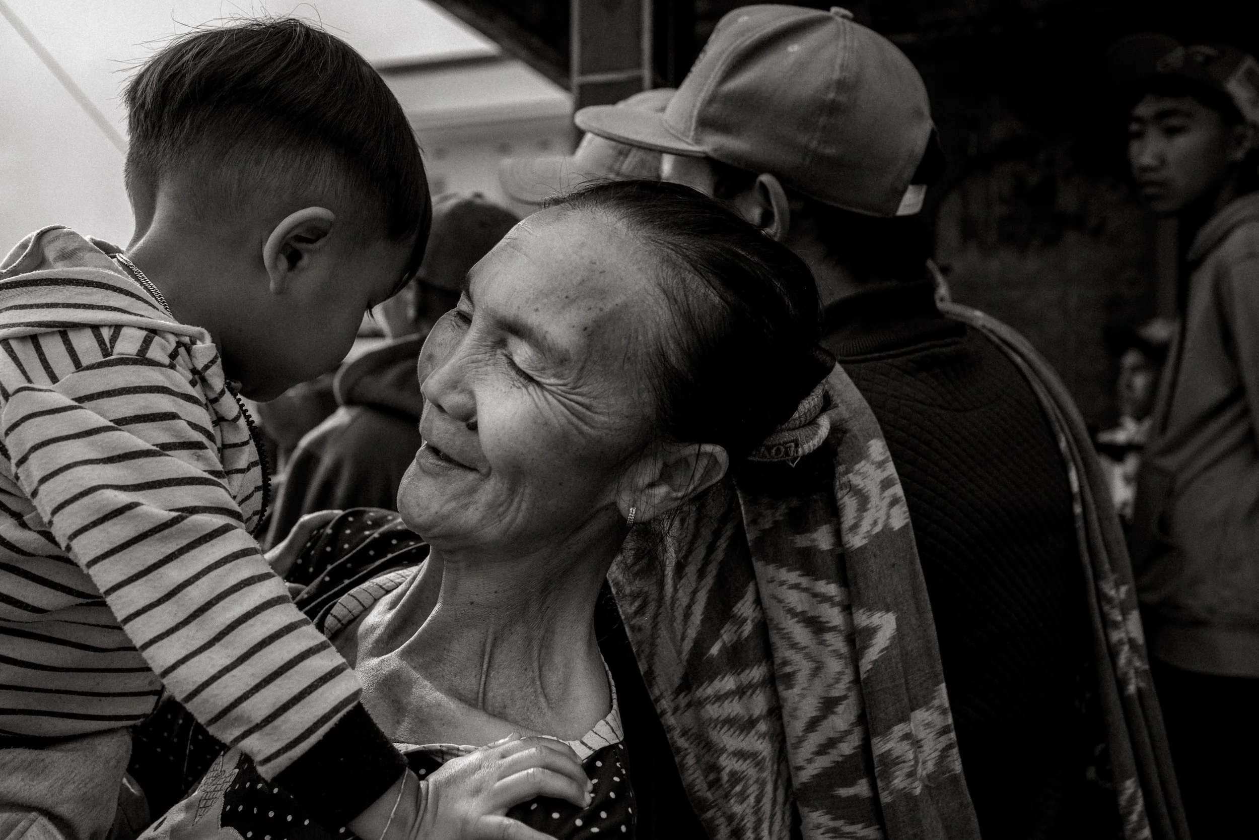 A woman smiles with a child from the edge of the square where spectators gather. She stands among a crowd of men. Ngadas, East Java, Indonesia. 21 August 2025.