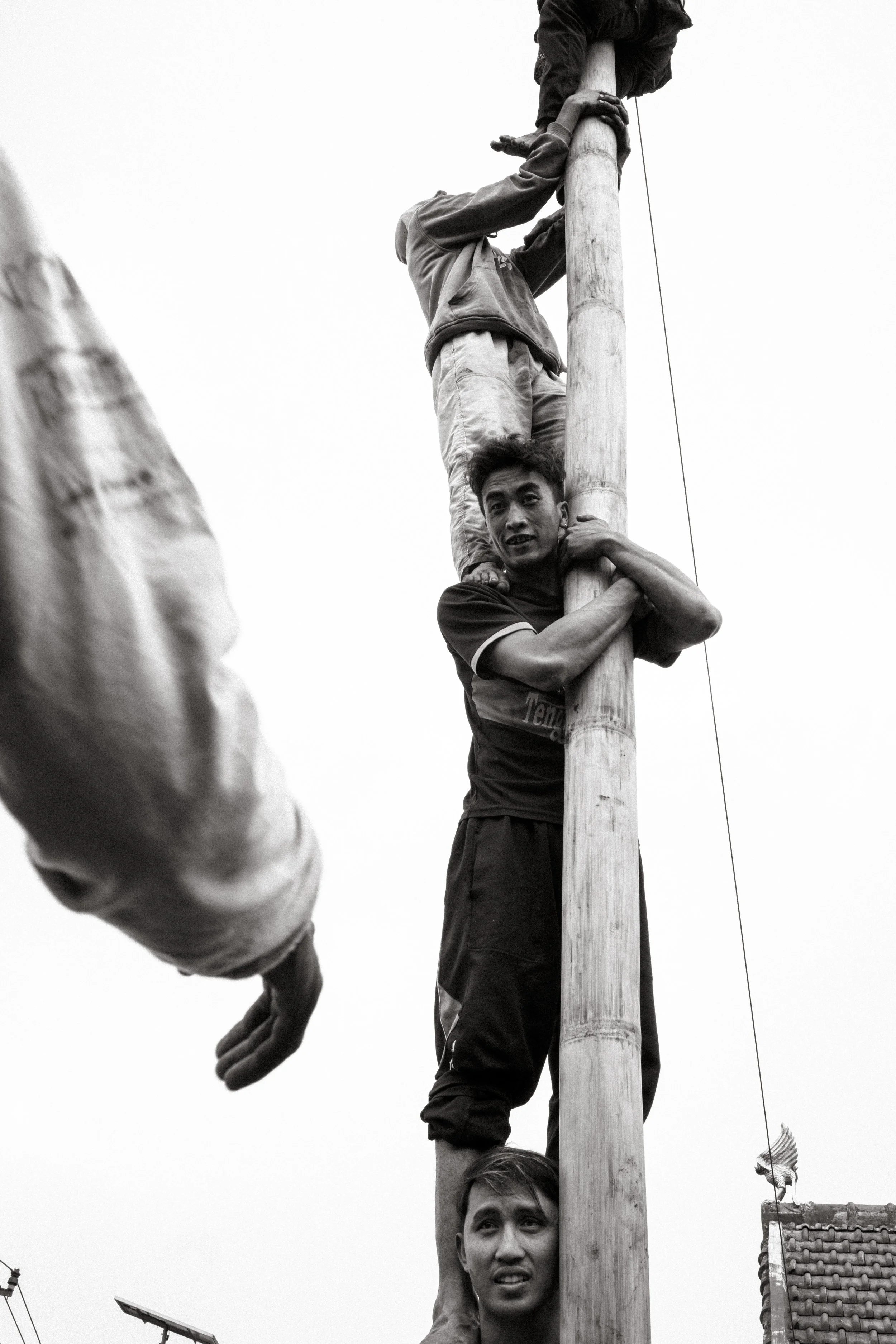 Three young men are working together to climb and hold onto a tall wooden utility pole. One man is at the bottom, supporting the pole with a focused expression. The second man is in the middle, gripping the pole with his arms crossed, and the third m