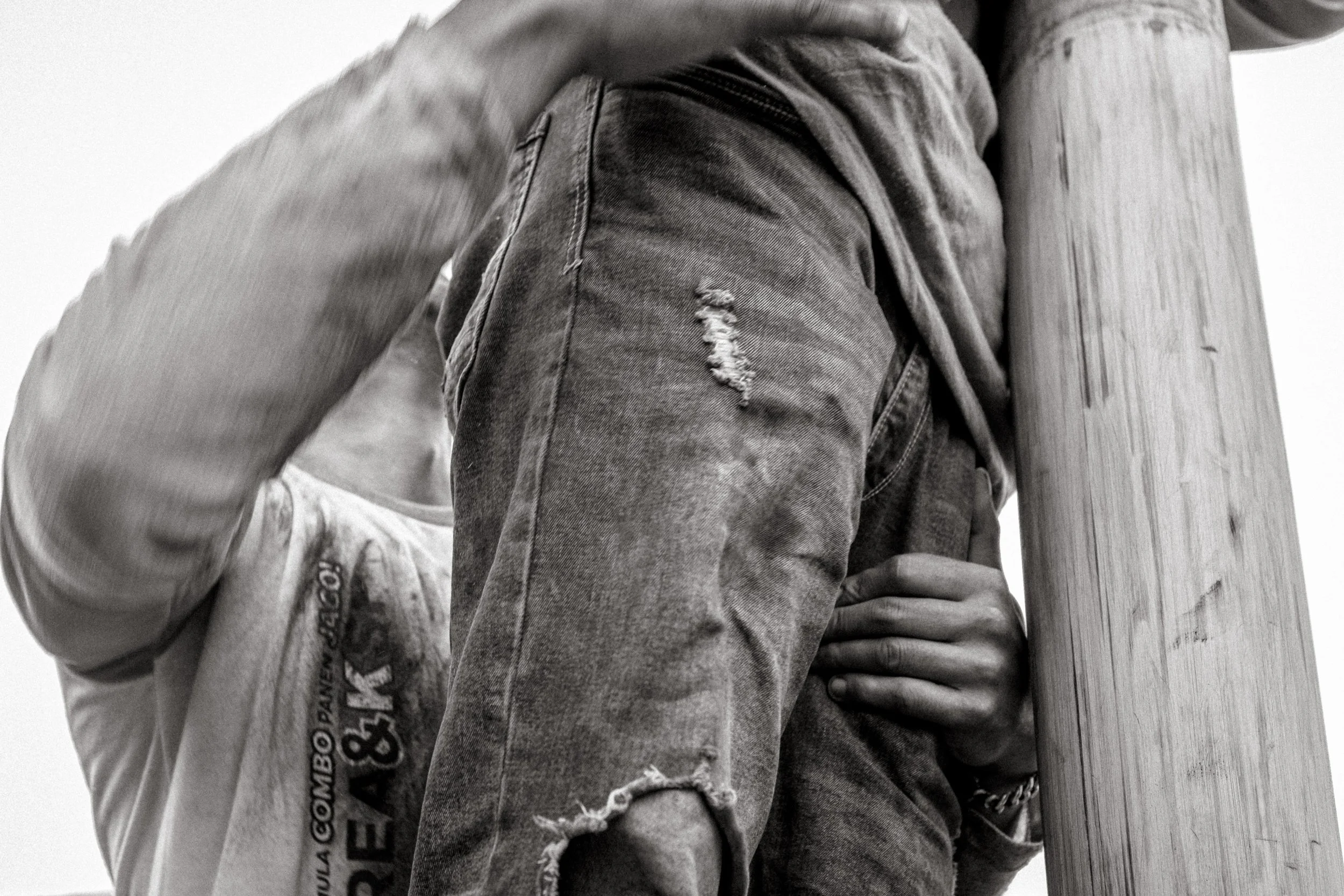 A climber is supported by his legs and waist as he climbs higher. Bodies stack and grip the pole, dirt and oil visible on their clothes. Ngadas, East Java, Indonesia. 21 August 2025.