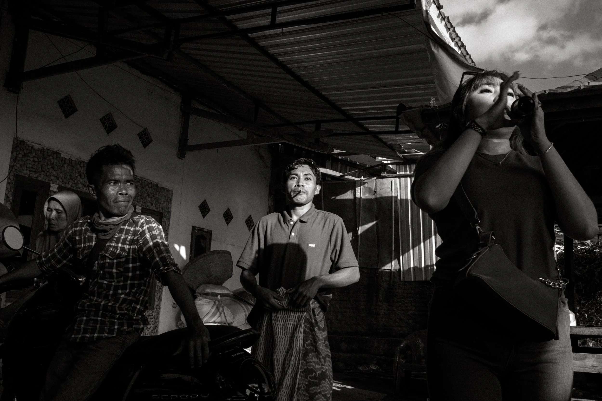A singer performs into a microphone beside a sound system while guests watch from shade