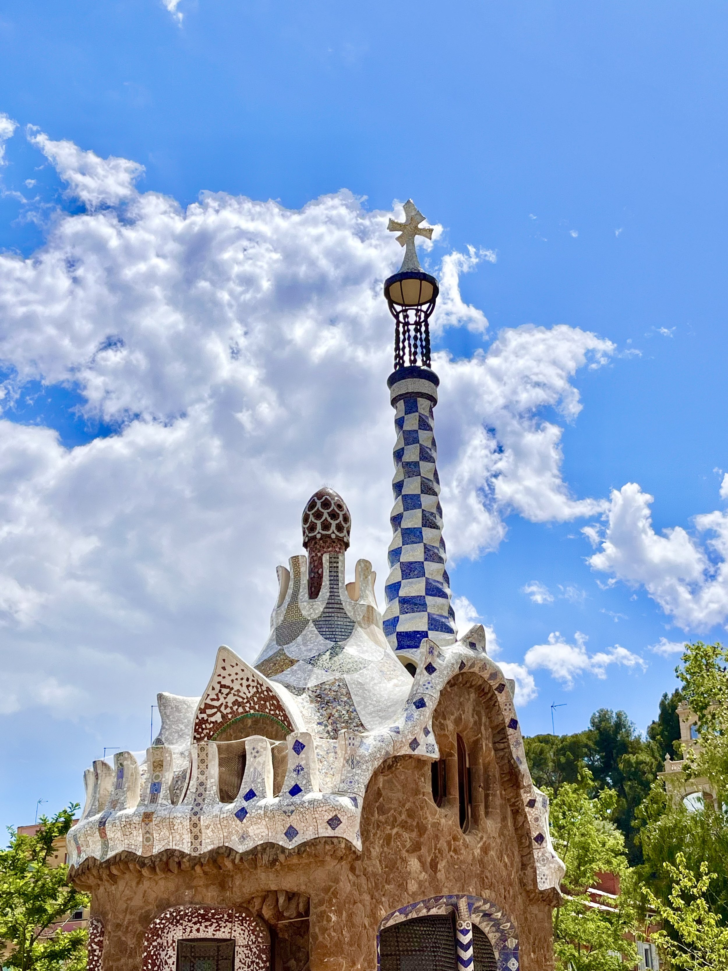 Colorful architectural structure with curved, mosaic-like surfaces, topped with a cross, set against a bright blue sky with clouds and green trees in the background.
