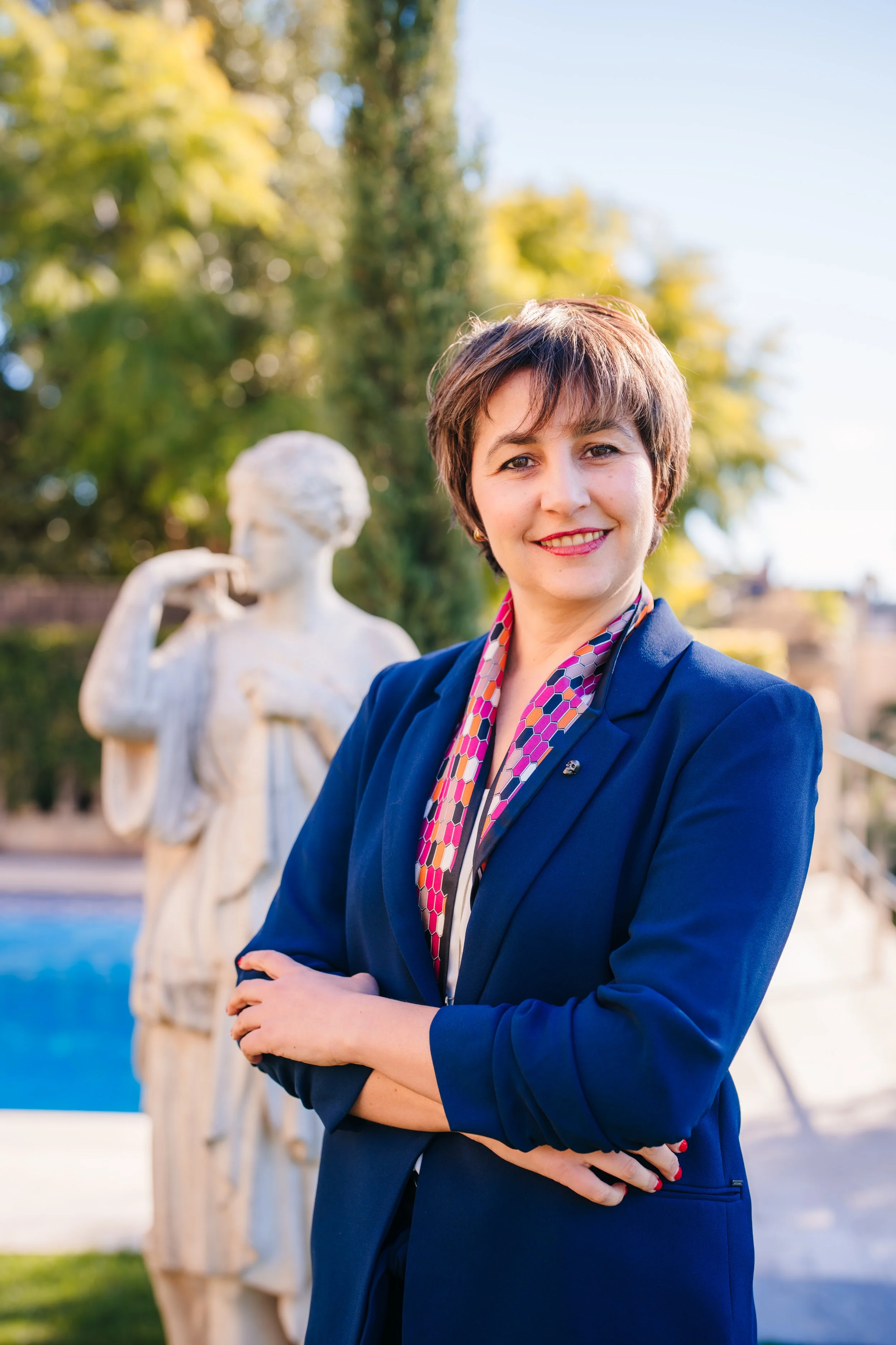 A woman with short brown hair and red lipstick standing outdoors, smiling, with crossed arms, in front of a white classical statue of a woman, and green trees in the background, during daytime.