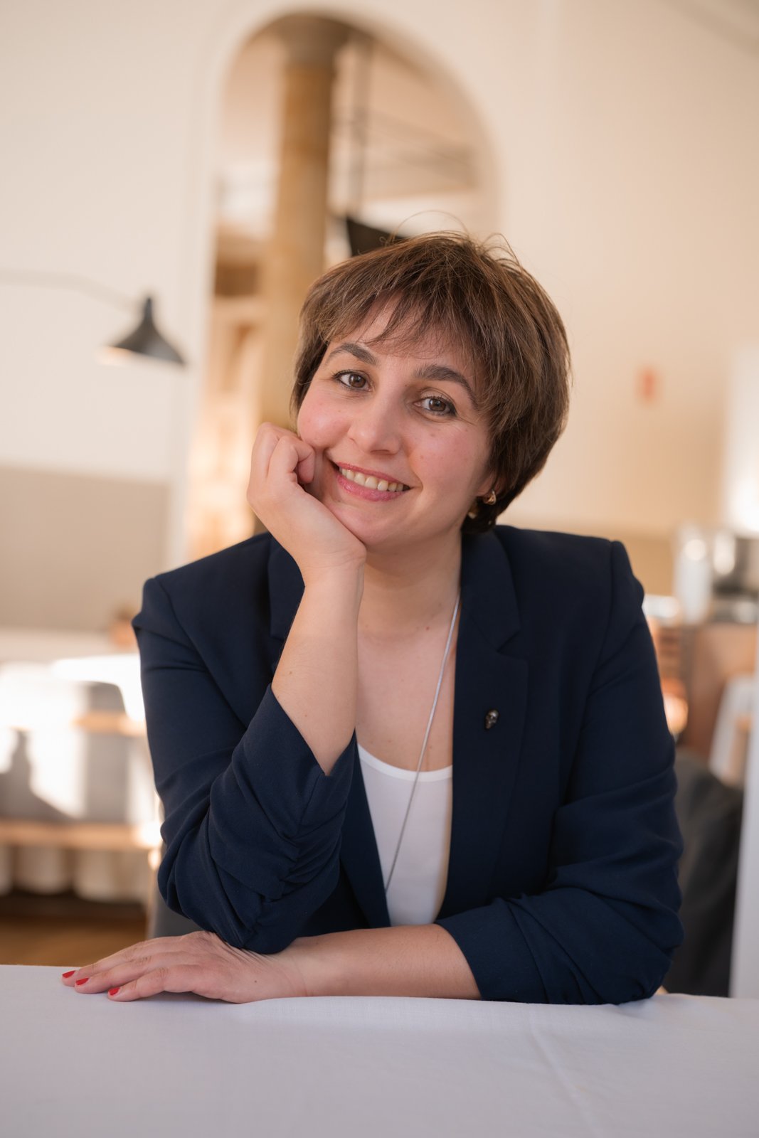 A woman with short brown hair smiling, resting her chin on her hand, wearing a dark blazer and white top, sitting at a table in an indoor setting with warm lighting.