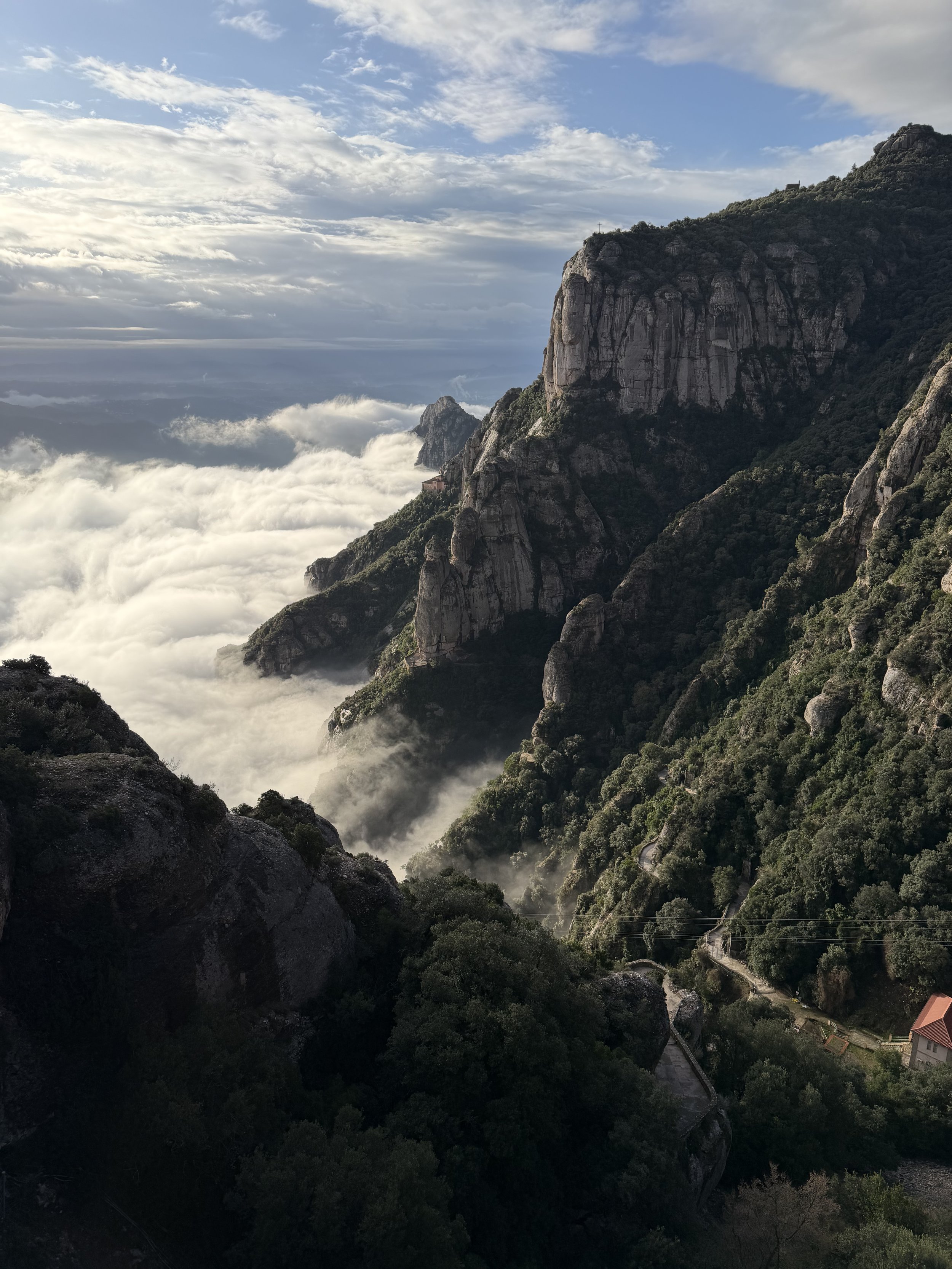 Mountain landscape with rocky peaks, green vegetation, and clouds below the mountains, with a partly cloudy sky overhead.