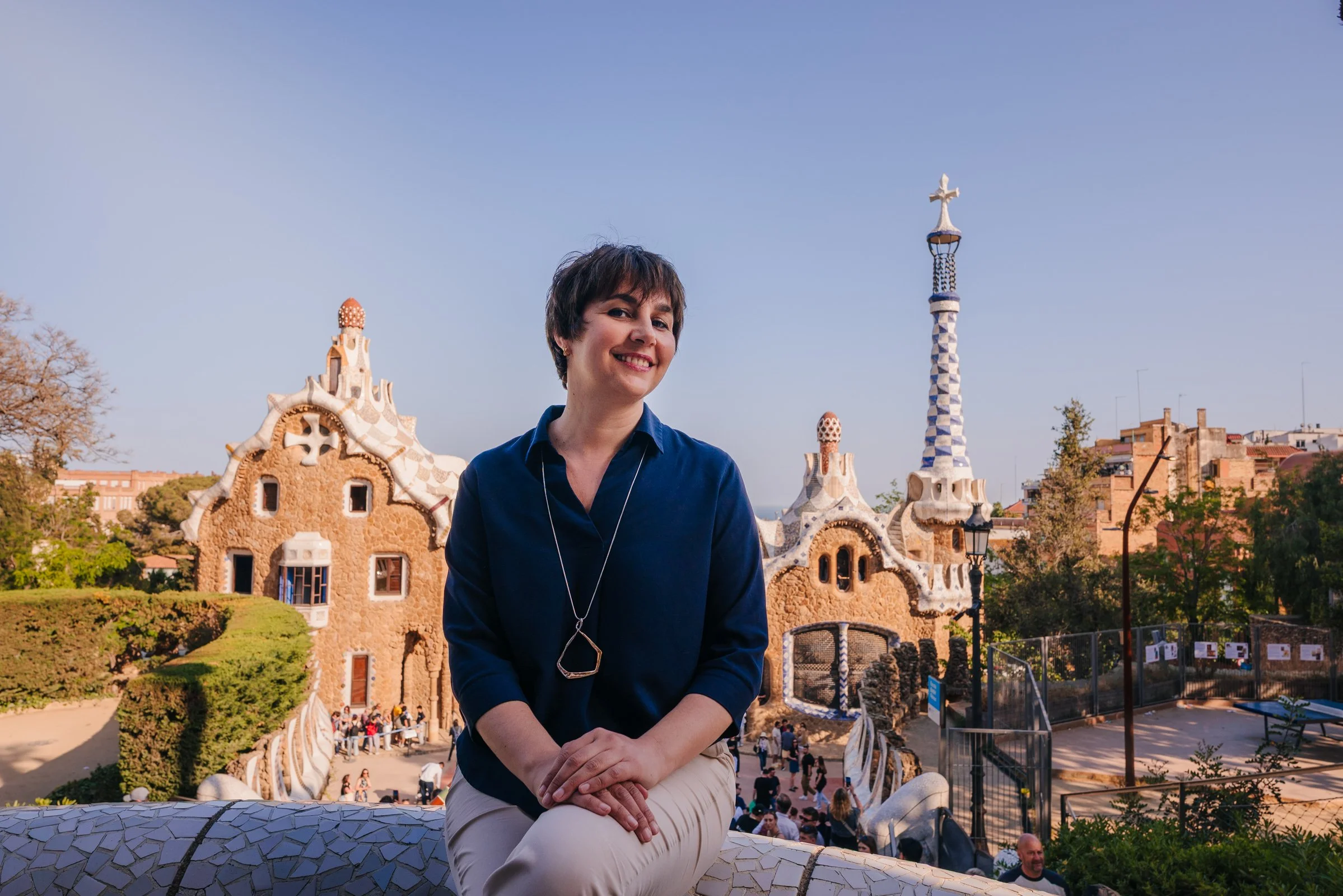 Woman with short dark hair smiling, sitting outdoors on a mosaic bench, with Park Güell architecture and a blue sky in Barcelona, Spain, in the background.
