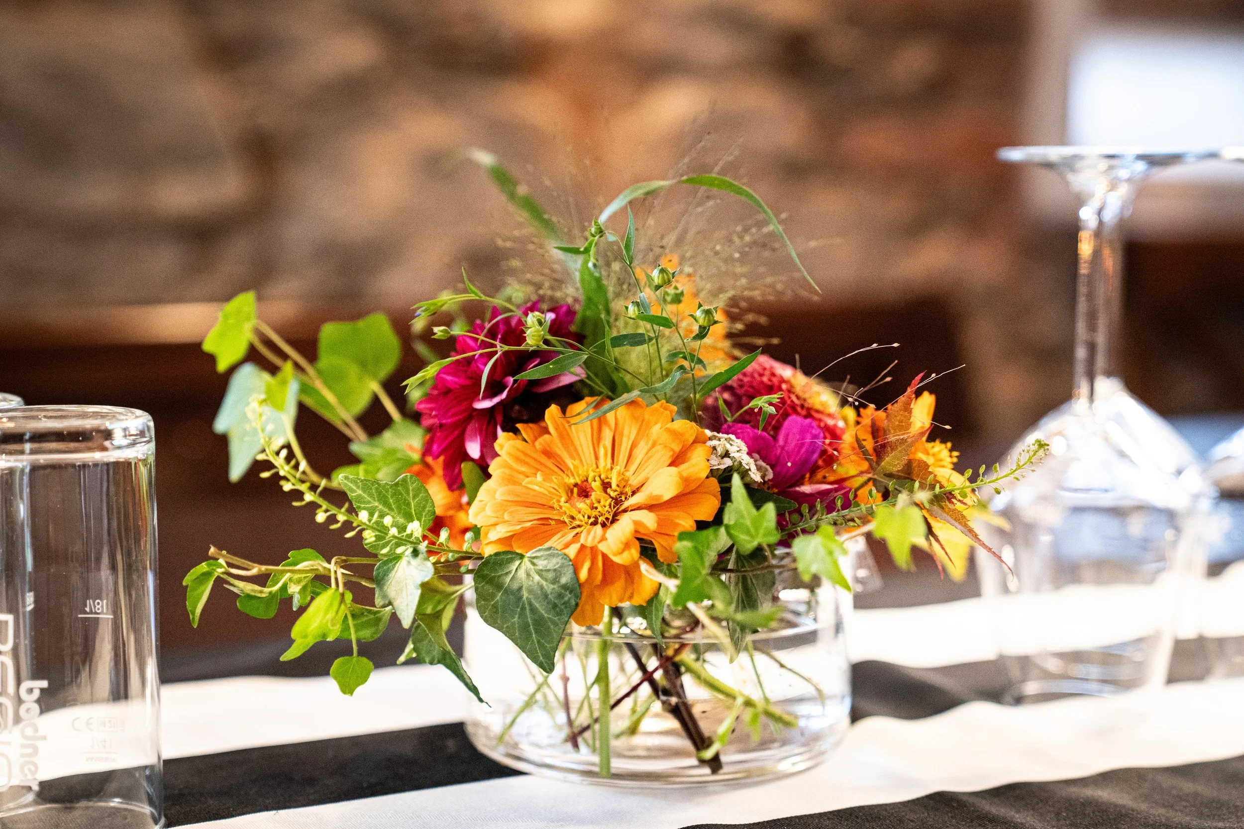 Blumenstrauß in einer Glasvase auf einem Tisch, mit Wasser gefüllt, umgeben von Wasser gläsern, im Hintergrund eine Steinwand.