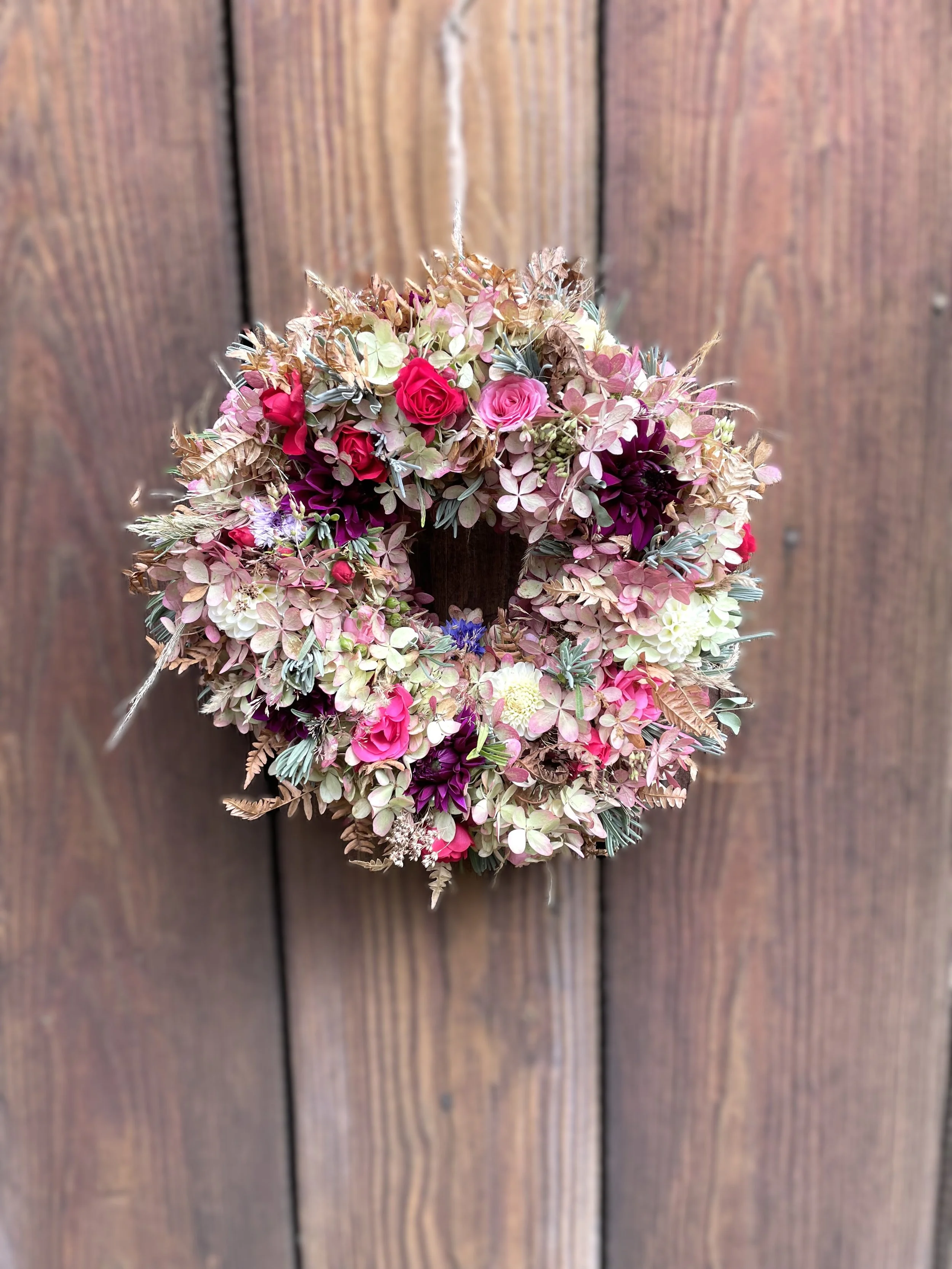 Blumenkranz aus verschiedenen pinken, roten und weißen Blumen hängt an einer Holzwand.