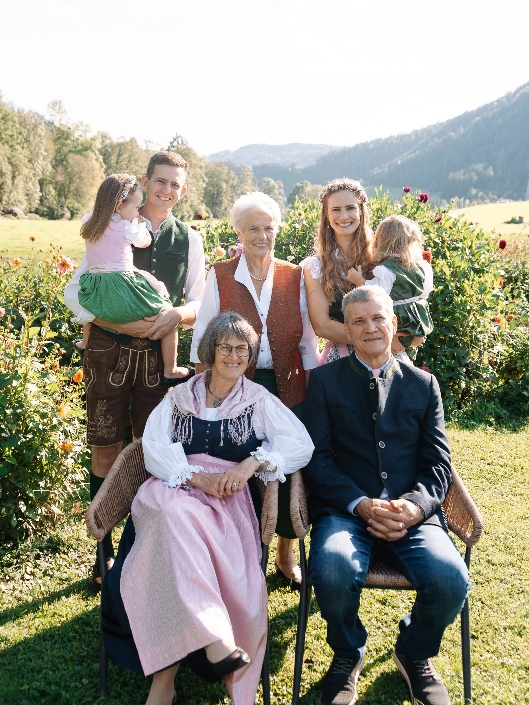 Eine Gruppe von sieben Menschen, drei ältere Frauen, zwei jüngere Frauen, zwei jüngere Männer und ein kleines Mädchen, steht im Freien vor einem Holzgebäude mit herbstlichen Blättern auf dem Boden.