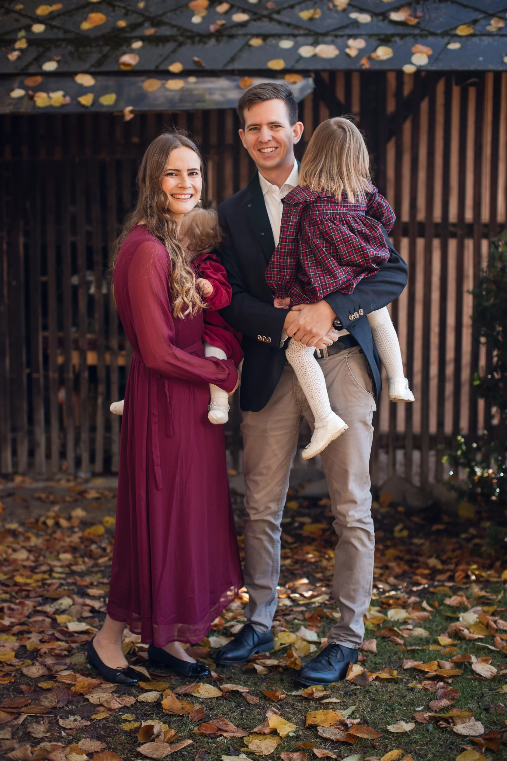 Eine Familie mit zwei Kindern bei herbstlichem Spaziergang, lachend vor einem Holzhaus, umgeben von gefallenen Blättern.