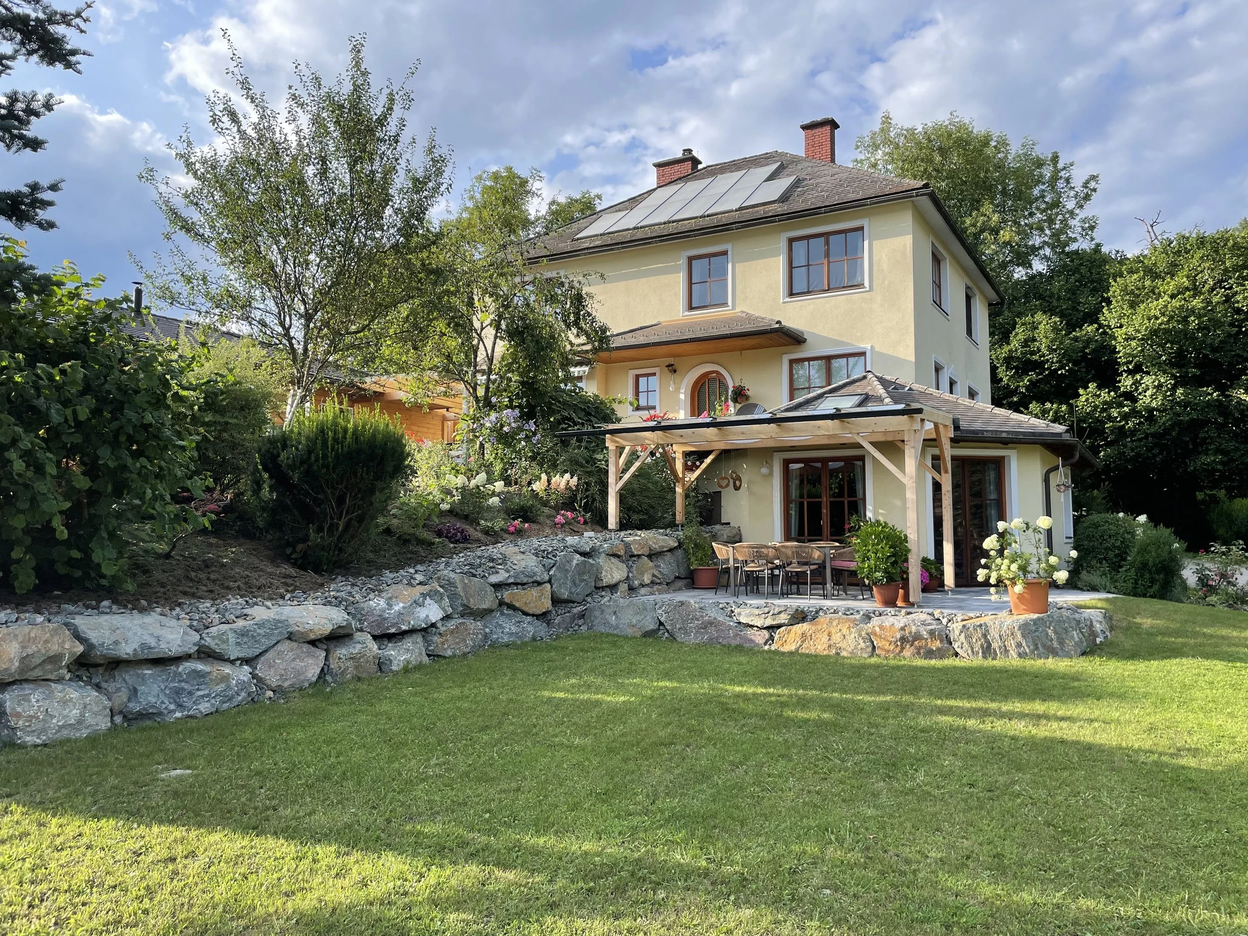 Ein zweistöckiges Haus mit einem gepflegten Garten und einer Terrasse im Freien, umgeben von Bäumen und Sträuchern, bei sonnigem Himmel.