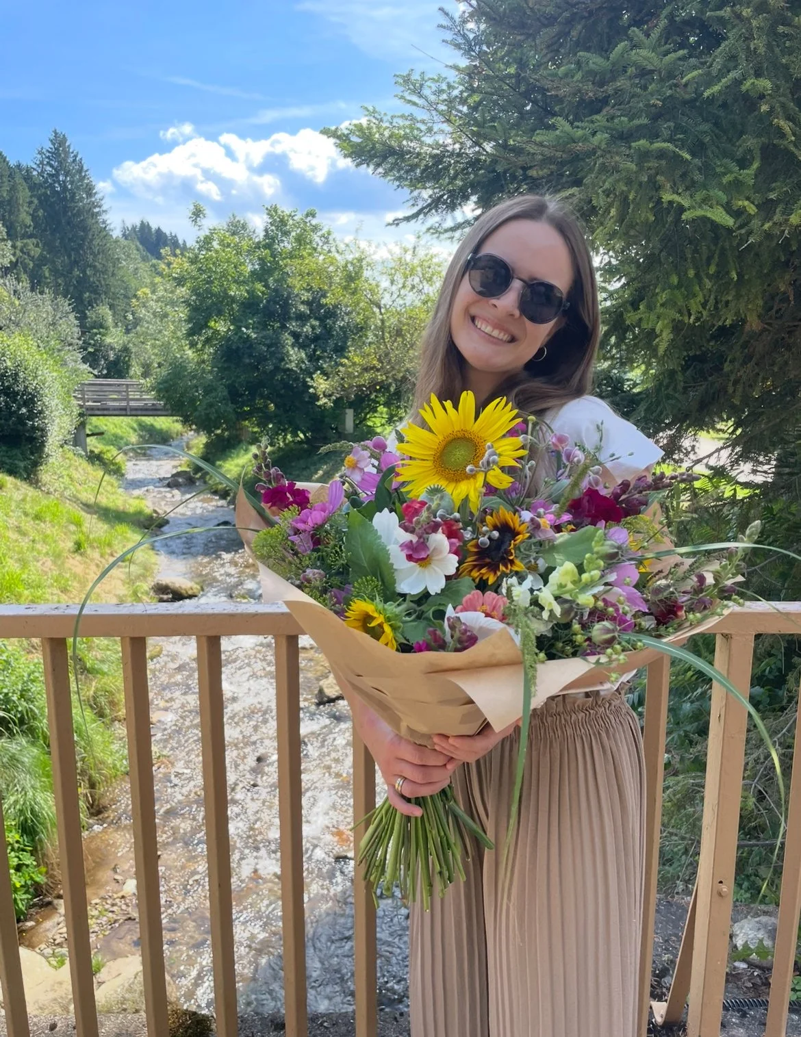 Junge Frau mit Sonnenbrille hält Blumenstrauß vor einem Fluss im Grünen bei Sonnenschein.