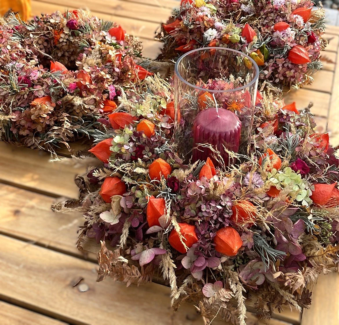 Herbstliche Blumenkränze mit roten Lampions, Kerzen und Trockenblumen auf einem Holztisch.