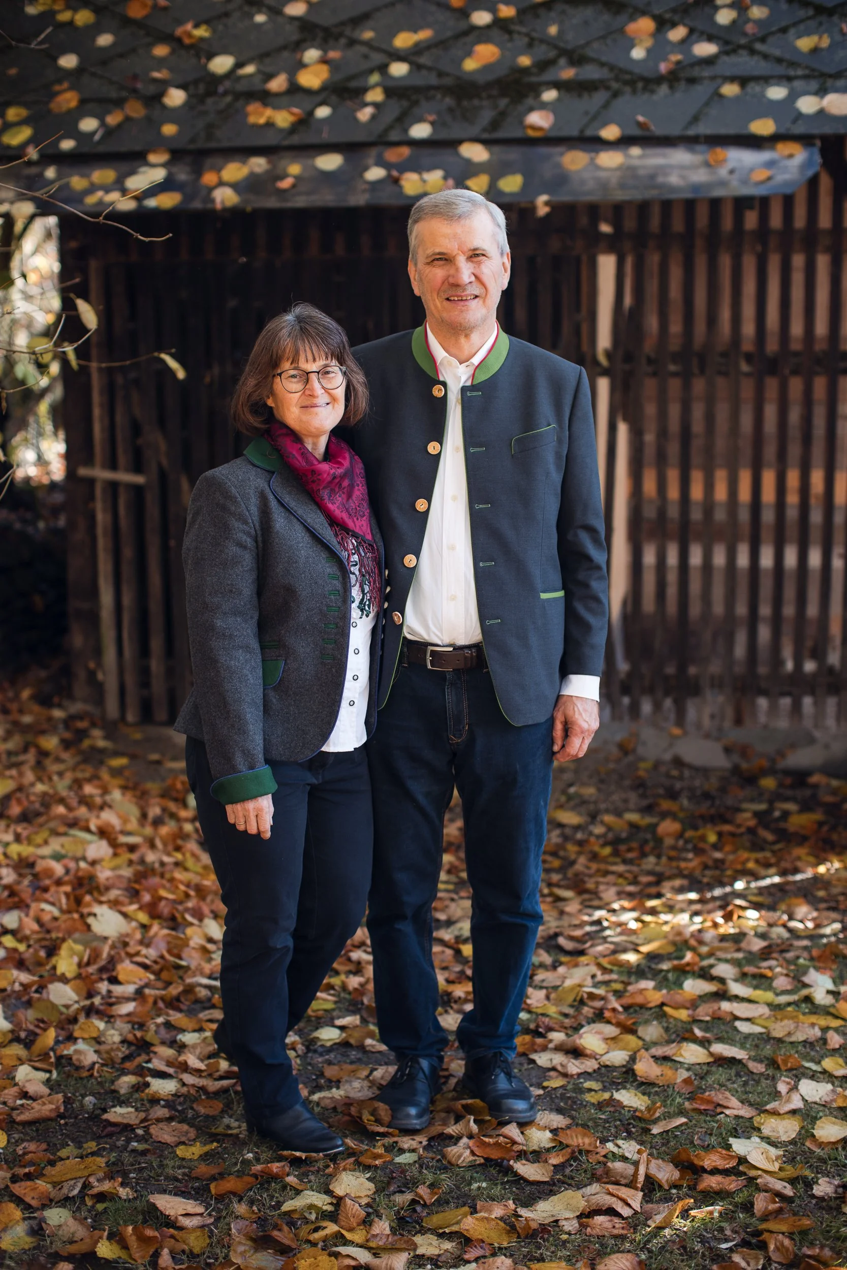 Ein älteres Paar steht im Freien vor einem Holzschuppen, umgeben von Laub im Herbst. Beide tragen traditionelle bayerische Trachtenjacken und lächeln in die Kamera.