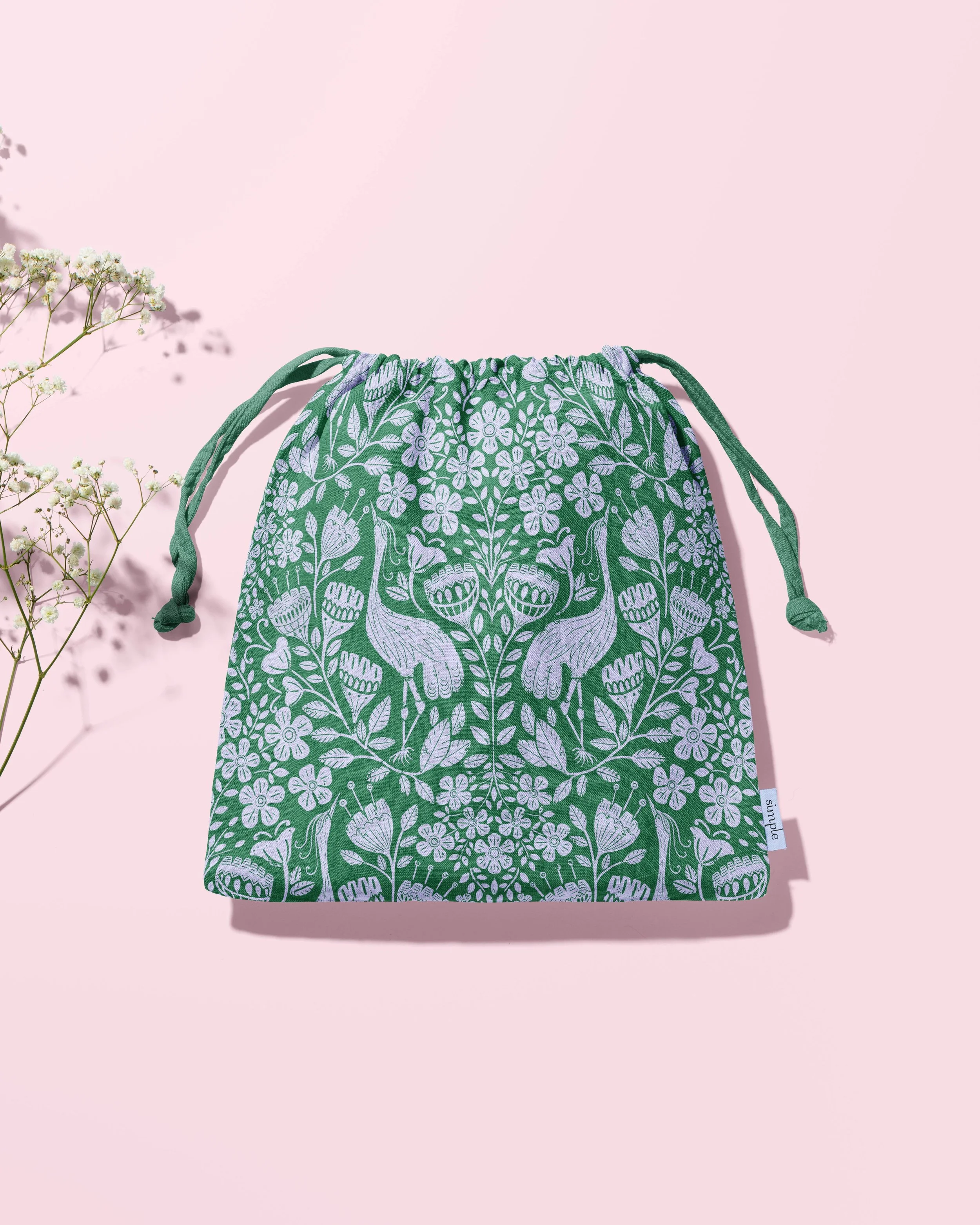 A green drawstring bag with white floral and animal print design, placed on a pink background next to some small white flowers.