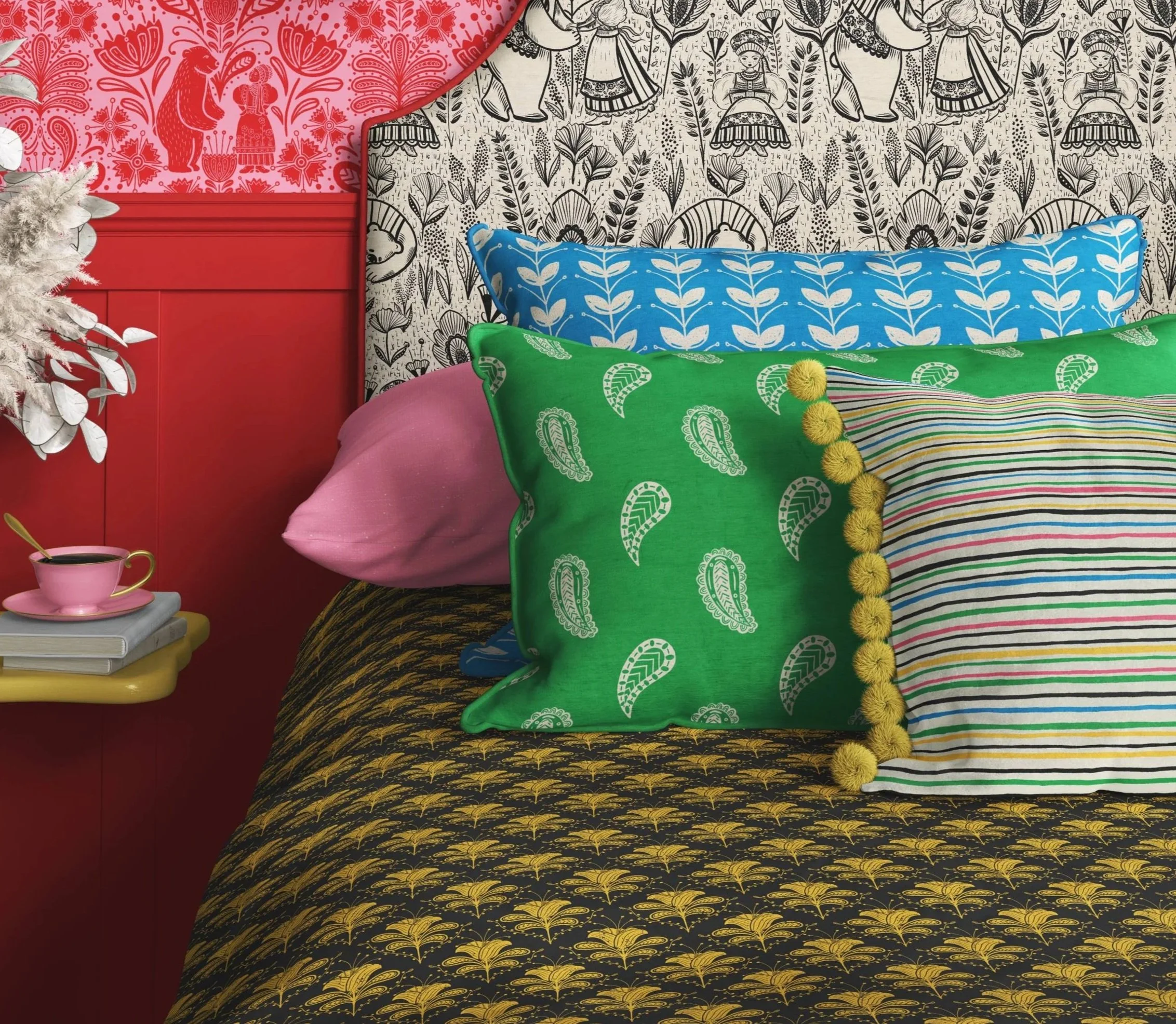 Colorful bedding with patchwork of floral and nature-inspired patterns, including a green pillow with yellow pom-poms, a blue pillow with white leaf motifs, a pink pillow, and a black pillow with gold designs. A small pink cup and saucer are on a yellow side table with books.
