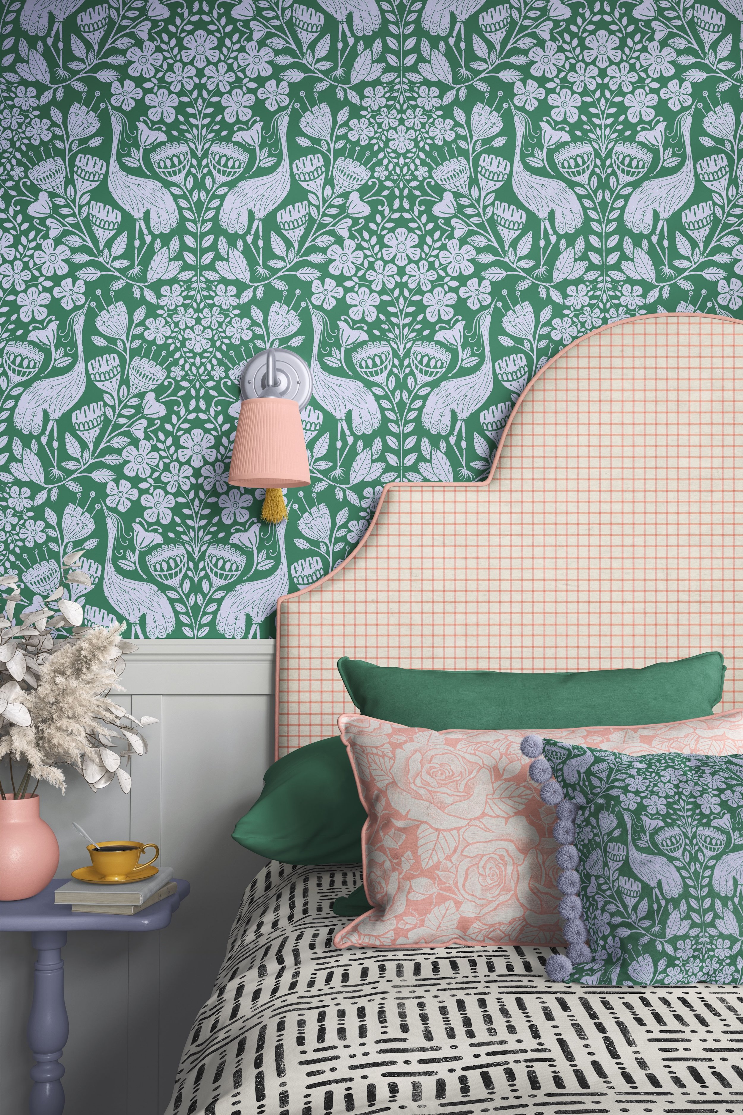 A bedroom corner with a bed featuring a pink and white checkered headboard, decorated with green, pink, and patterned pillows. To the left, a small side table holds a pink vase with dried flowers and a yellow cup and saucer. The wall has green and wh