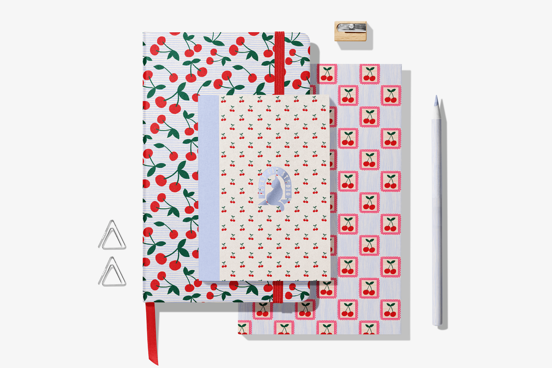 Flat lay of stationery with cherry themes, including patterned notebooks, gift tags, a paper clip, a pencil, and a stamp, all arranged on a white background.