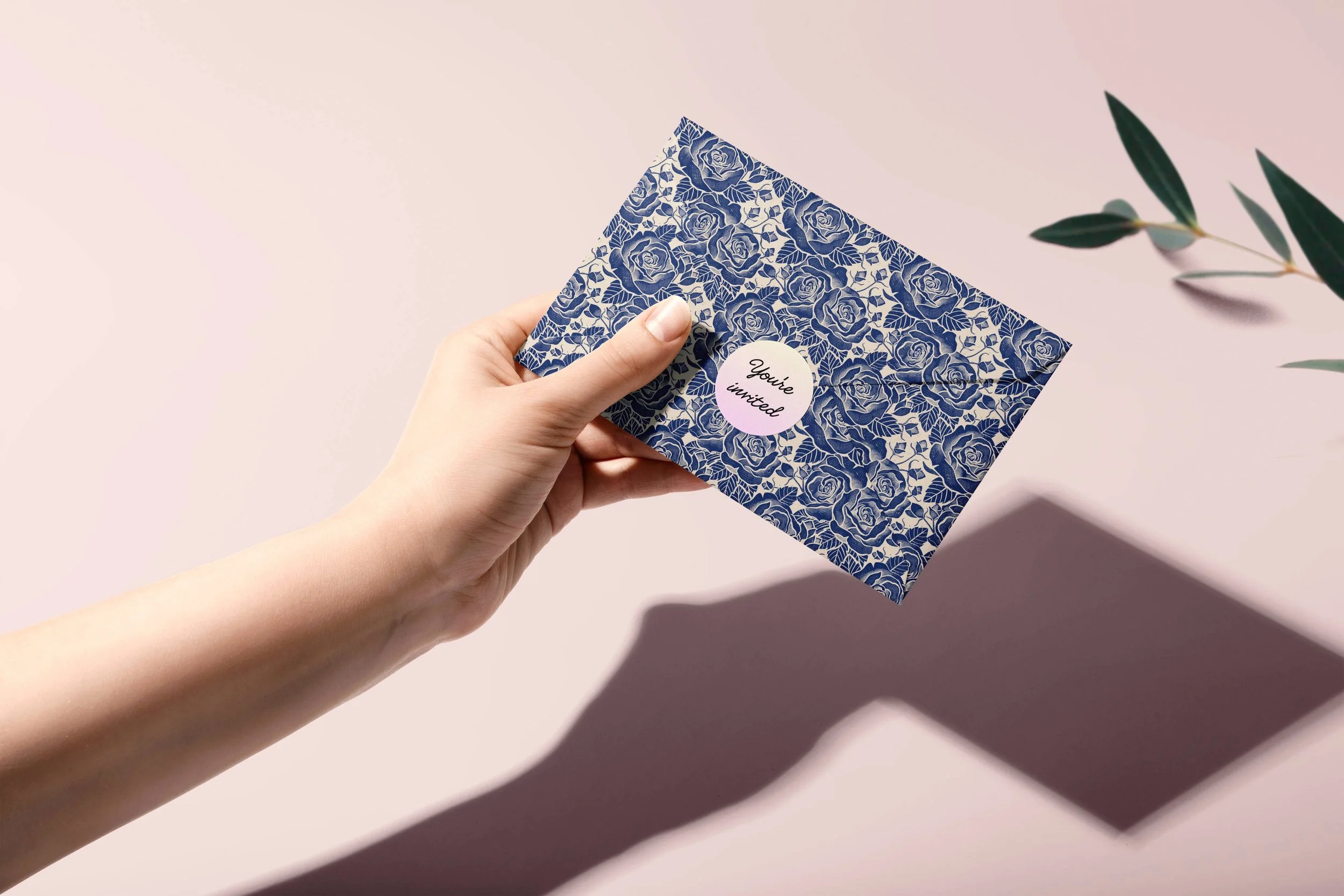 A person holding a blue floral patterned invitation card with the words 'You're invited' printed on a pink circular sticker. There are green leaves visible on the right side of the image and shadows cast on the pale pink background.